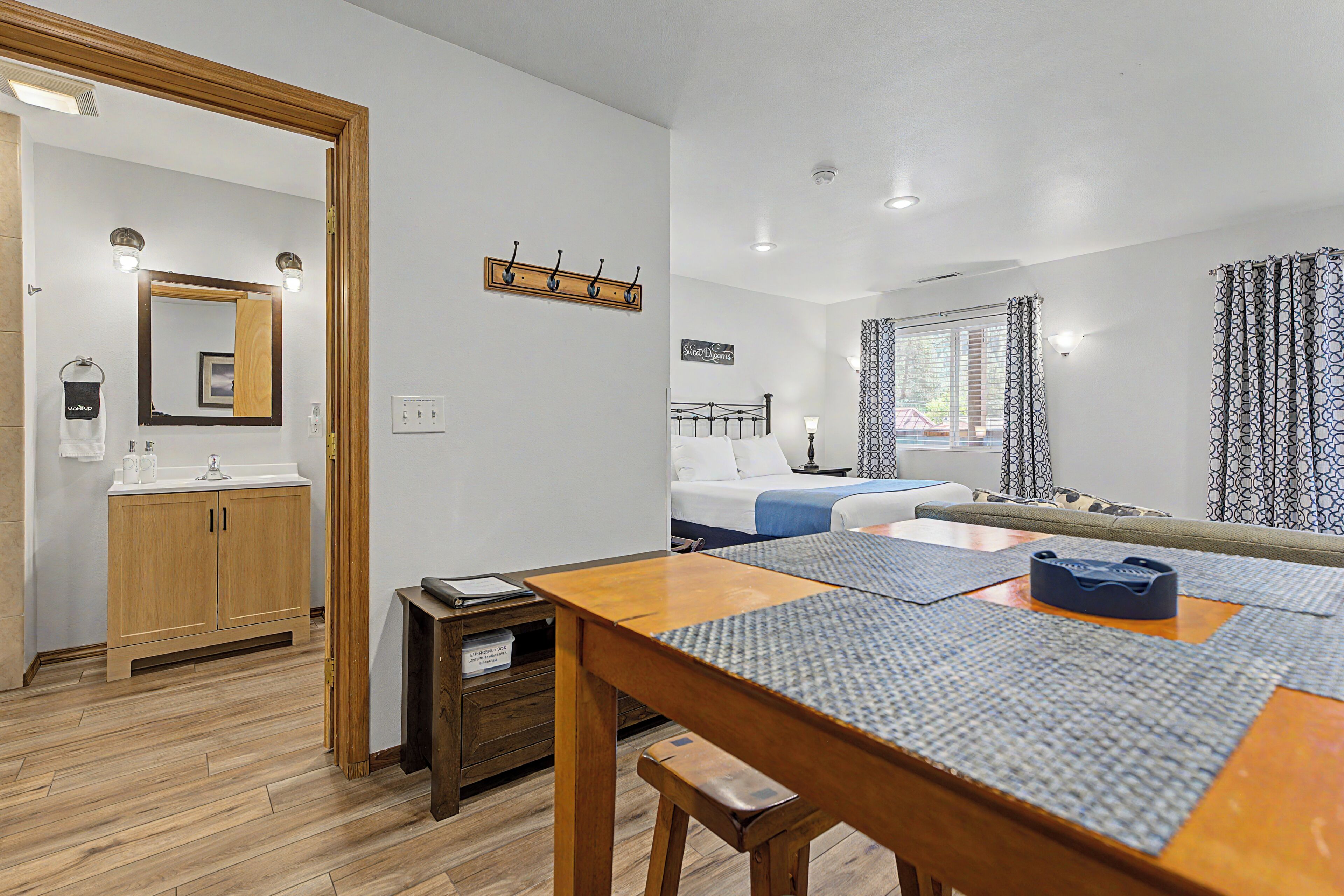 Quarto com mesa de madeira, cama, um banco, espelho e janela com cortinas.
