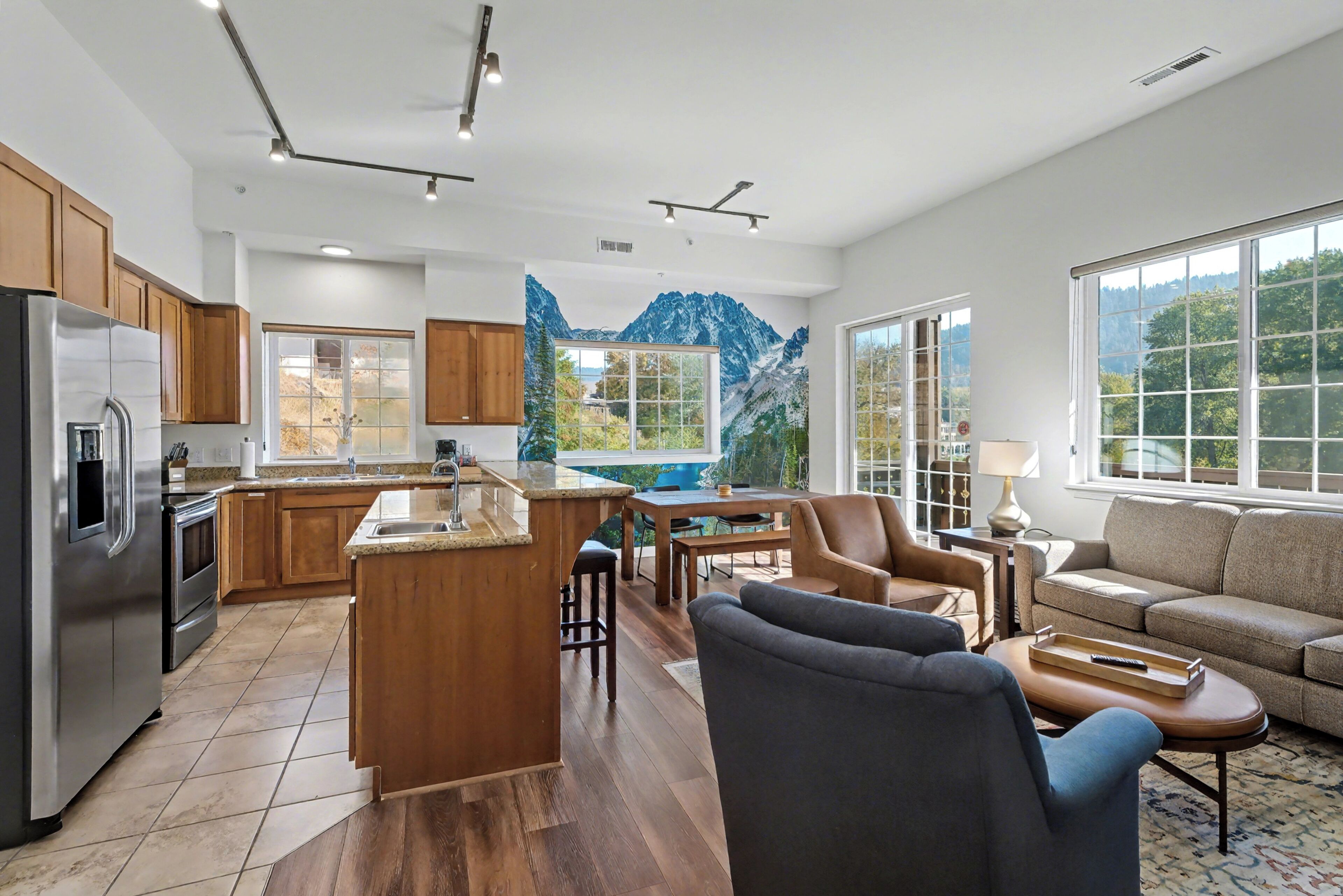 A modern kitchen with wooden cabinets, a stainless steel refrigerator, and a large island with a sink. There is a dining area with a table and chairs, and a sofa in the living room.