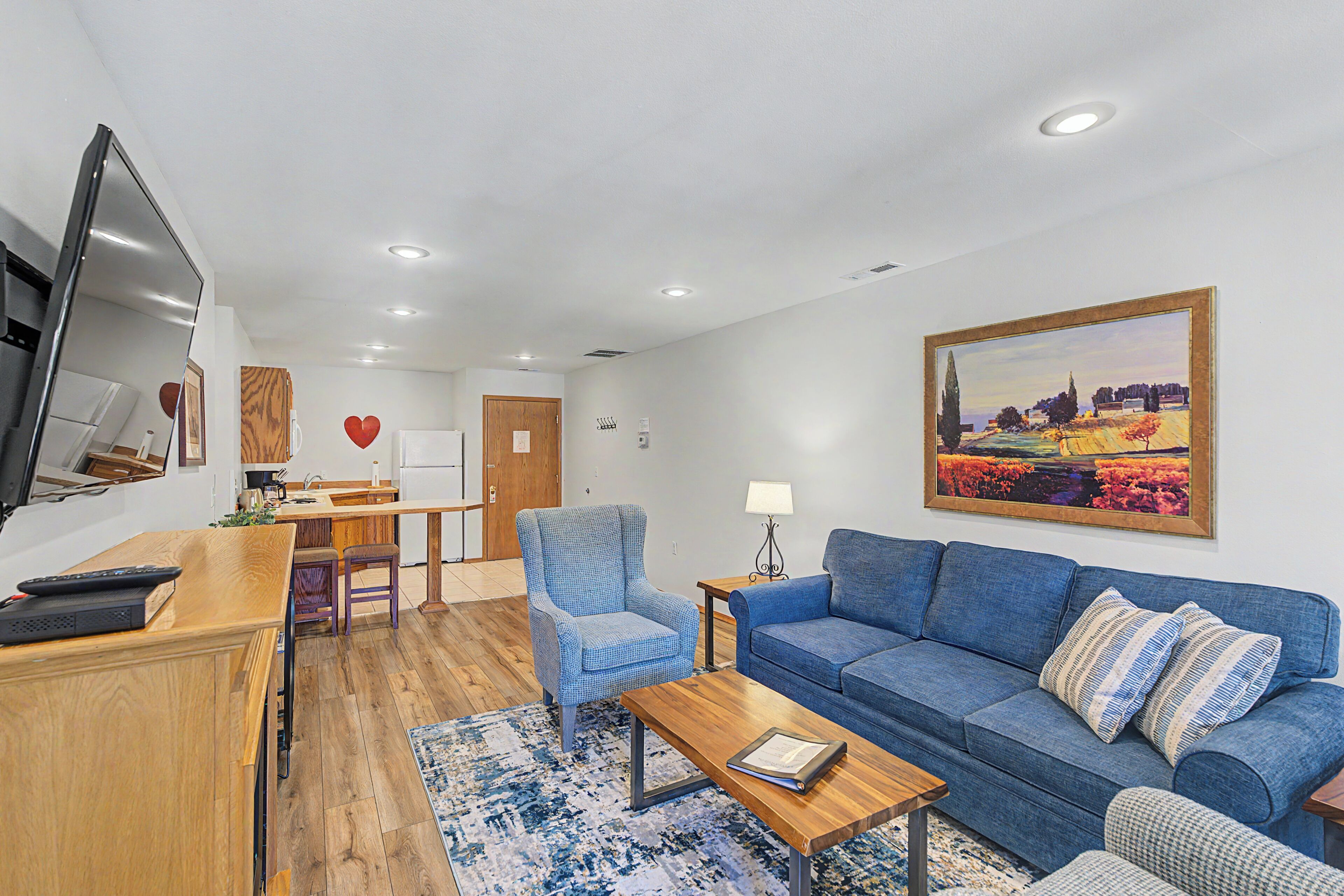 Un salon avec un canapé bleu, une table basse en bois, un fauteuil bleu, un téléviseur fixé au mur et un tableau accroché au mur.