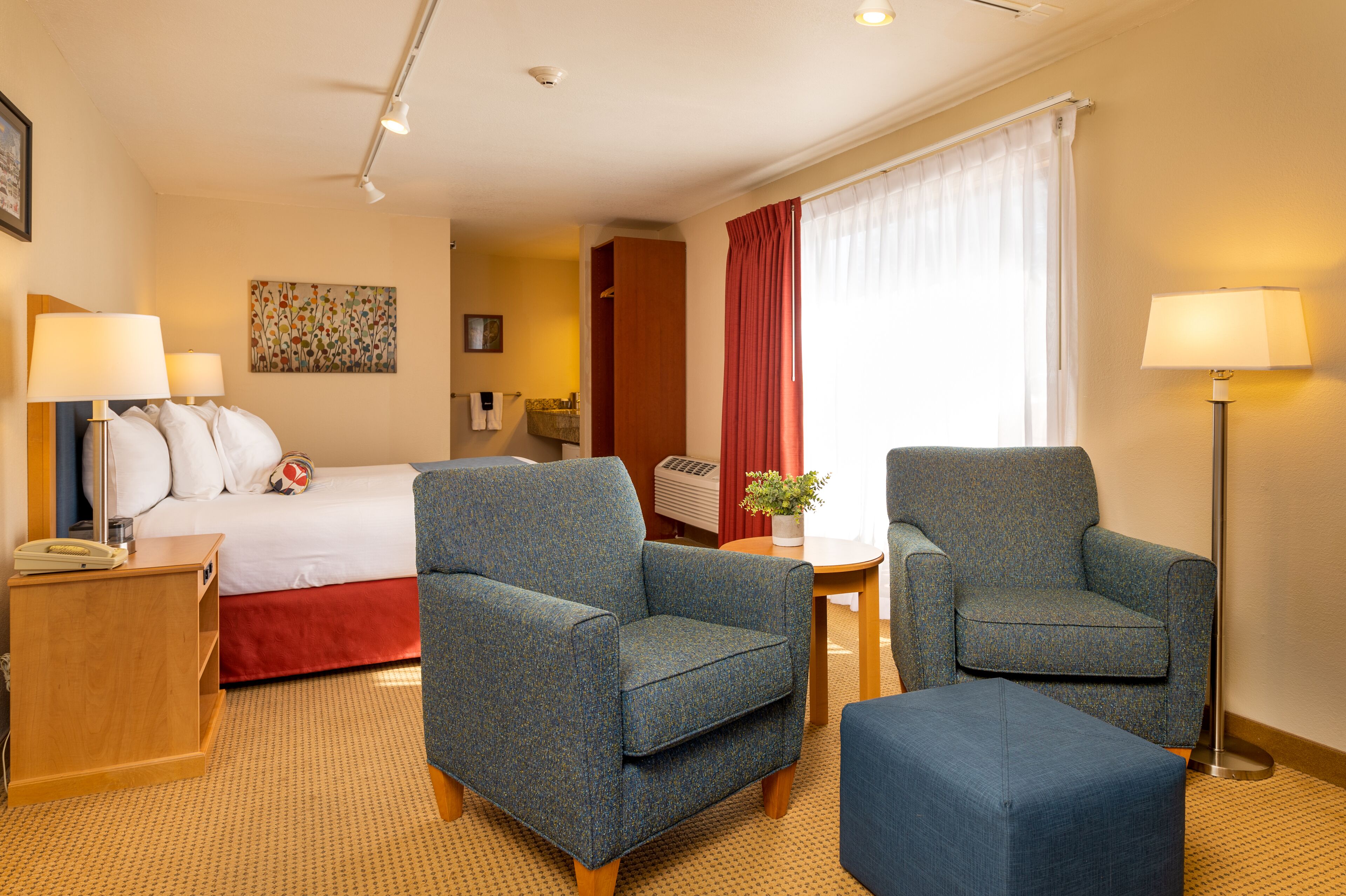 A hotel room with a bed, two armchairs, a side table, a lamp, a potted plant, and a window with curtains.