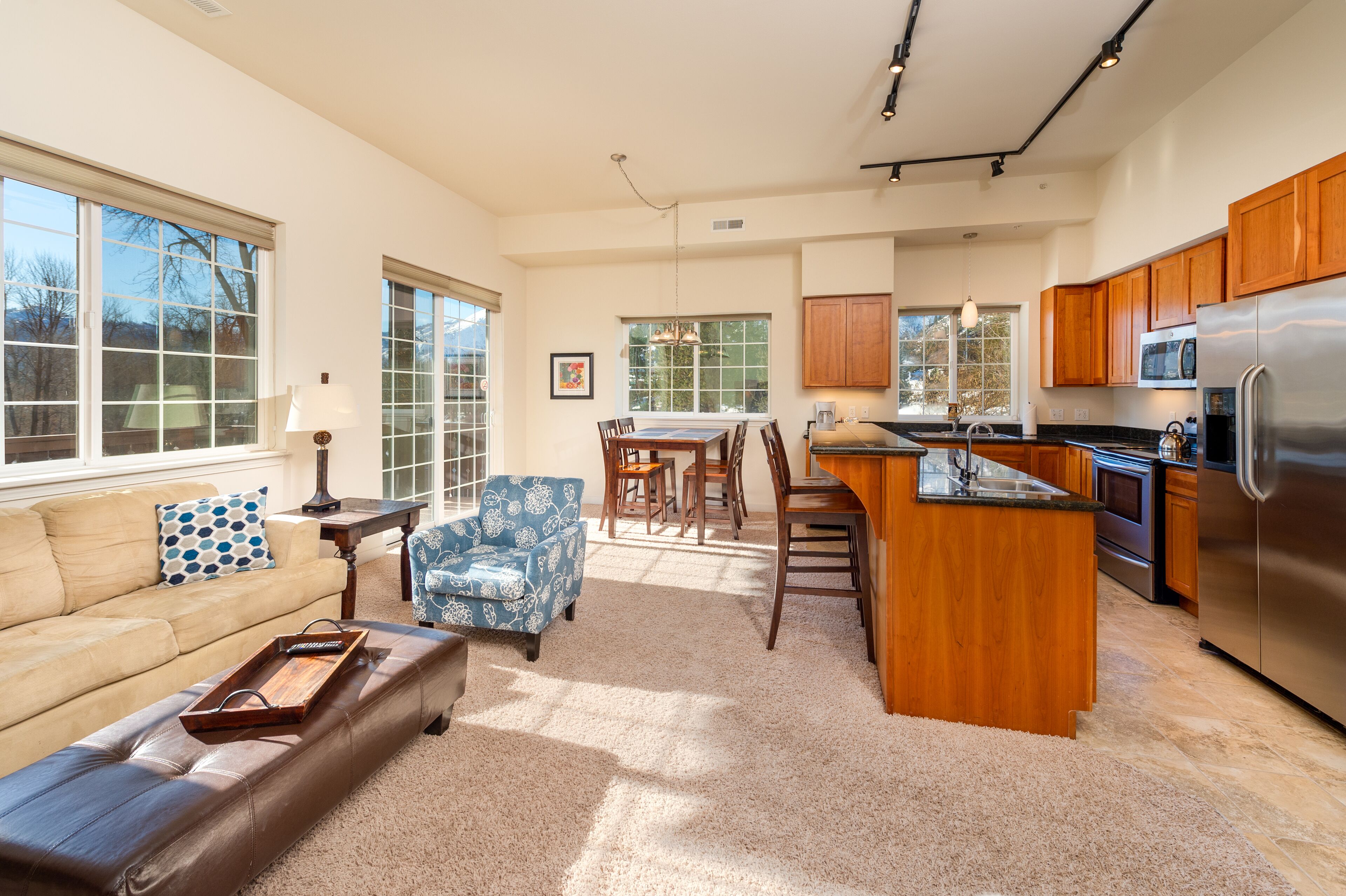 A spacious living room with a sofa, a patterned armchair, and a coffee table. A kitchen area with wooden cabinets and a stainless steel refrigerator is visible.