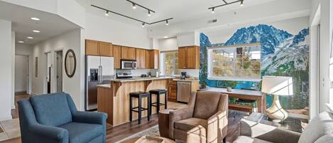 A modern living room with a blue armchair, a brown leather chair, and a wooden coffee table. A large window offers a view of mountains and a pool. The kitchen is visible in the background with wooden cabinets and a central island.