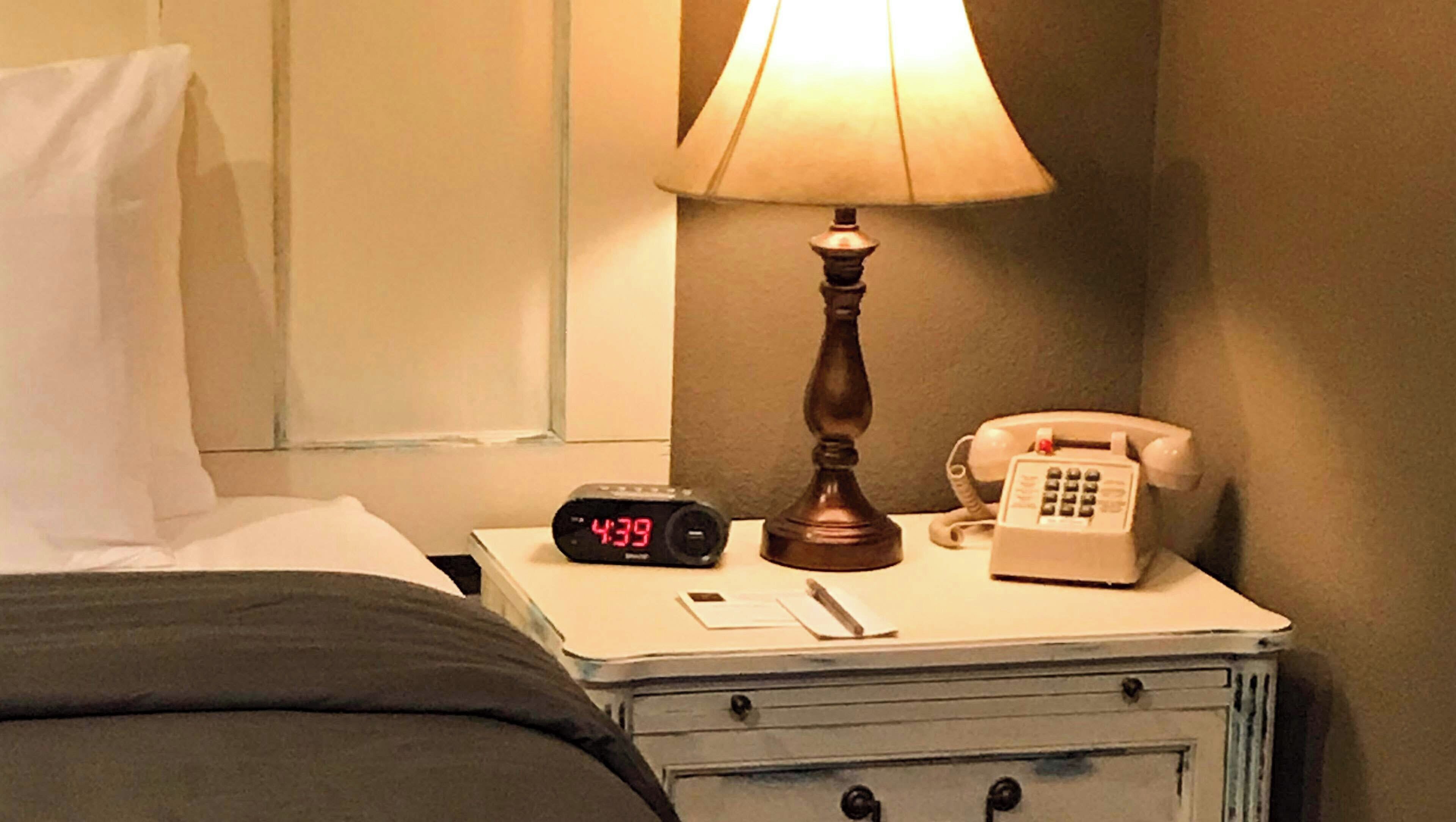 A bedside table with a lamp, a telephone, a digital clock, and a notepad.