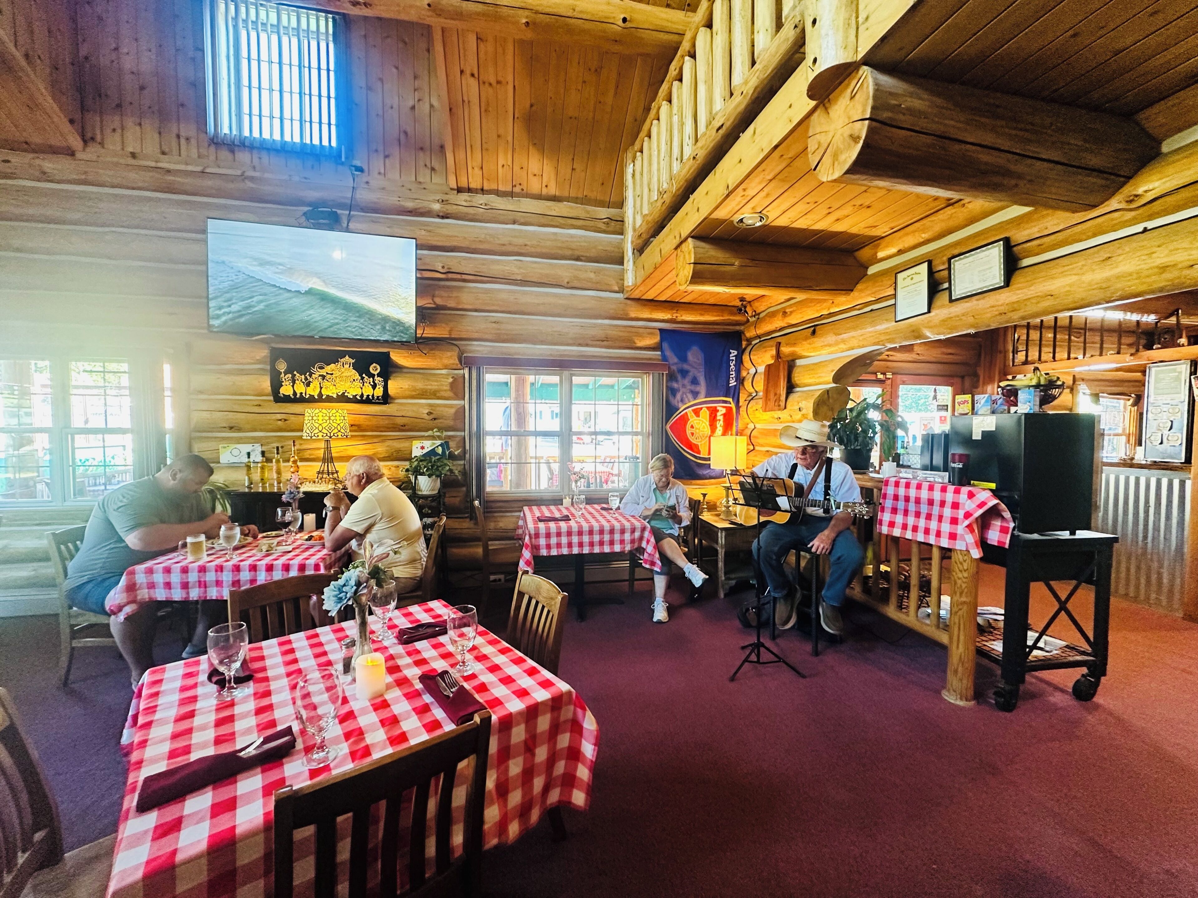 A rustic wooden interior with multiple tables covered in red and white checkered tablecloths, a television mounted on the wall, and a large window allowing natural light.