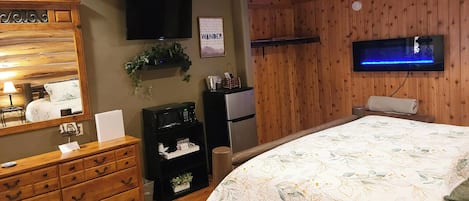 Un dormitorio con cabecera de madera, cama con colcha floral, cómoda, televisor y estante en la pared con una planta.