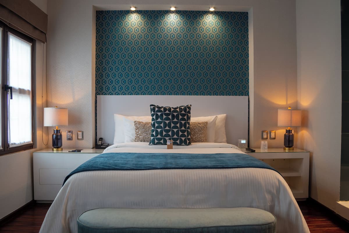 Una habitación de hotel moderna con una cama grande, dos mesitas de noche con lámparas, un papel tapiz decorativo y una ventana con persianas.
