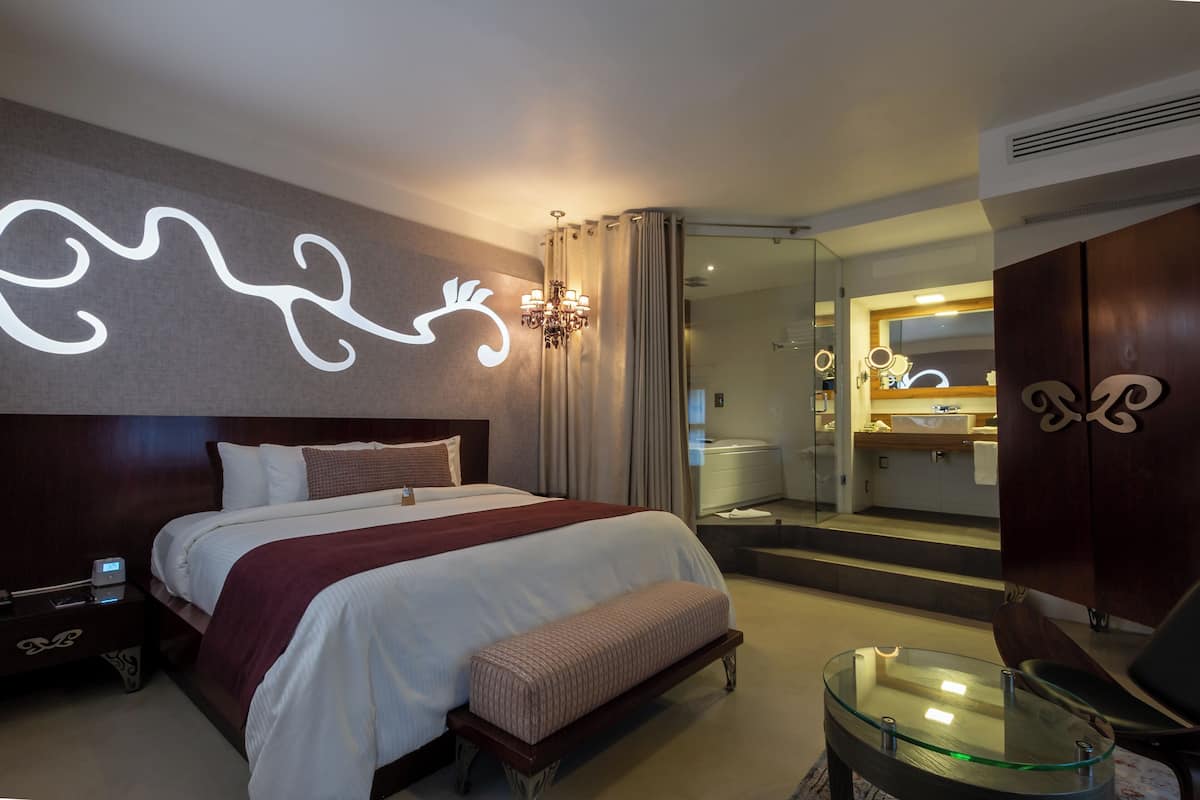 Una habitación de hotel moderna con una cama grande, una mesa de centro de vidrio, un banco y un baño visible a través de una puerta abierta.