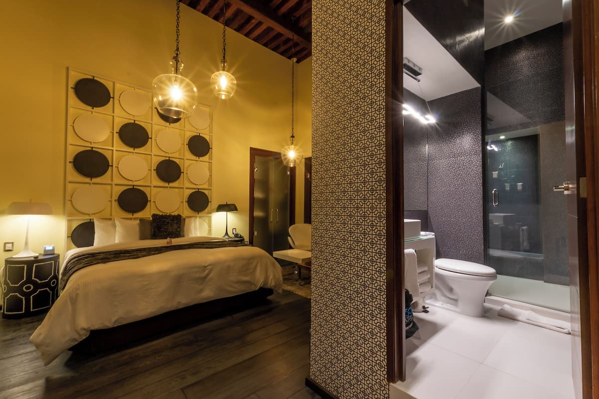 Habitación de hotel con cama, mesita de noche, lámpara de pared y baño con inodoro y ducha.