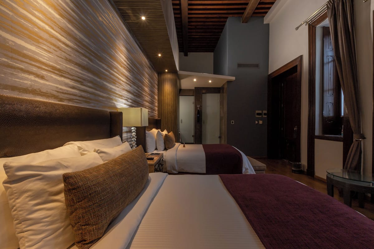 Habitación de hotel con una cama grande, cabecera con diseño texturizado, mesita de noche con lámpara y vista al pasillo.
