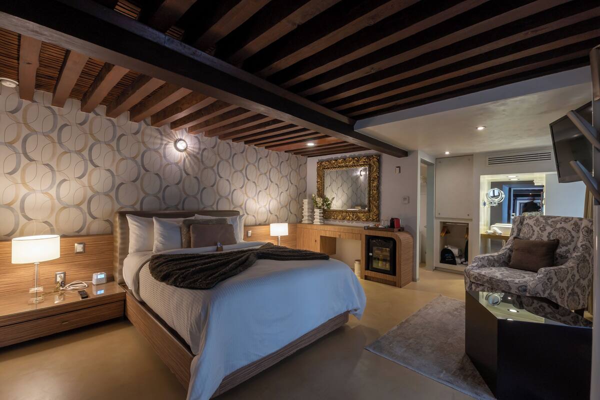 Un dormitorio moderno con una cama grande, lámparas de noche, cabecera de madera, un espejo, un televisor y un papel tapiz estampado.