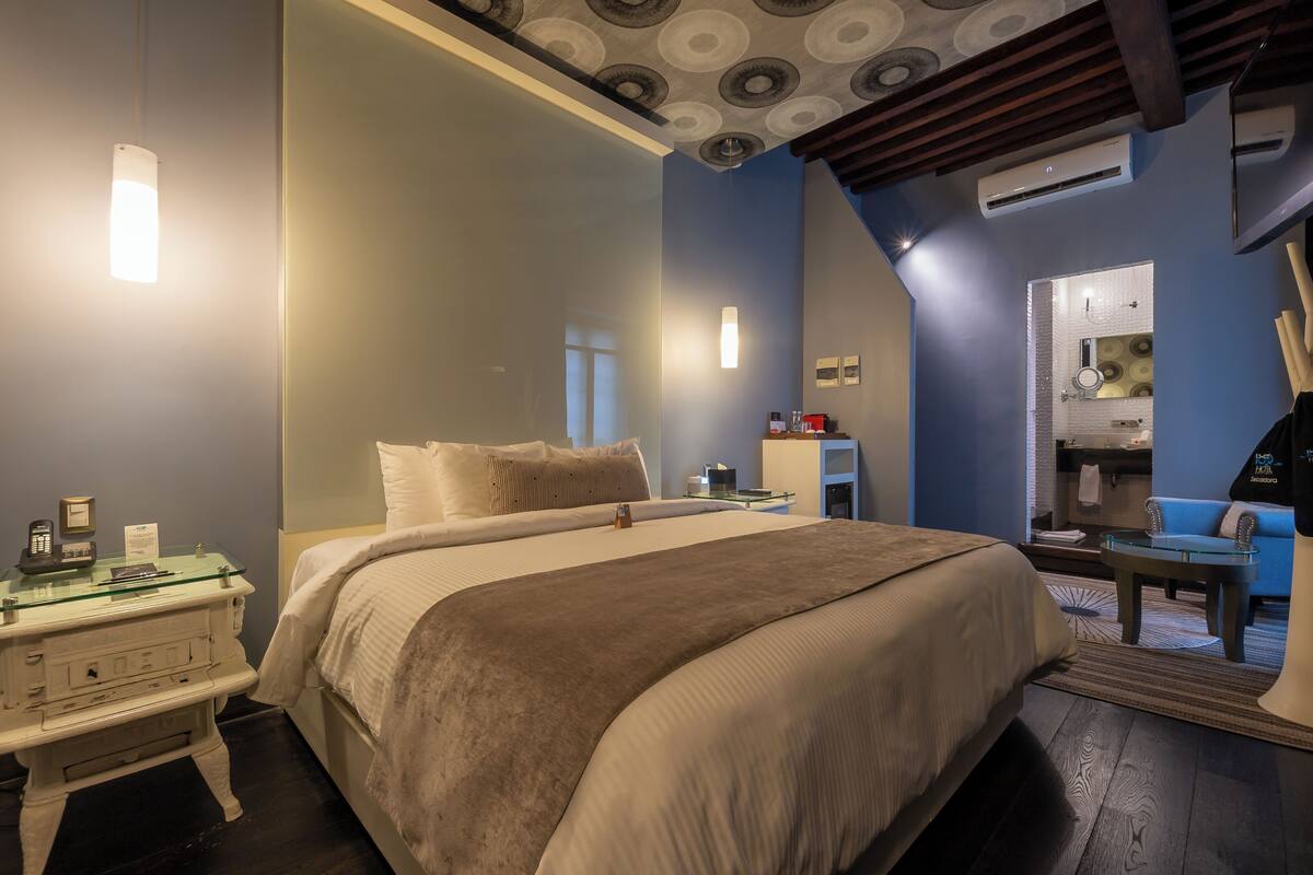 Una habitación de hotel moderna con una cama grande, mesitas de noche, una mesa de centro de vidrio y un área de descanso con un sofá y un sillón azules.