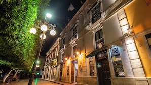 Une scène de rue nocturne, avec un immeuble doté d’un balcon et un magasin dont la vitrine expose des boissons en bouteille.