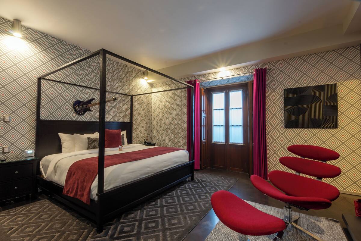Una cama con dosel, una manta roja, una mesita de noche con lámpara y un sillón rojo en una habitación con papel tapiz estampado y un ventanal con cortinas.