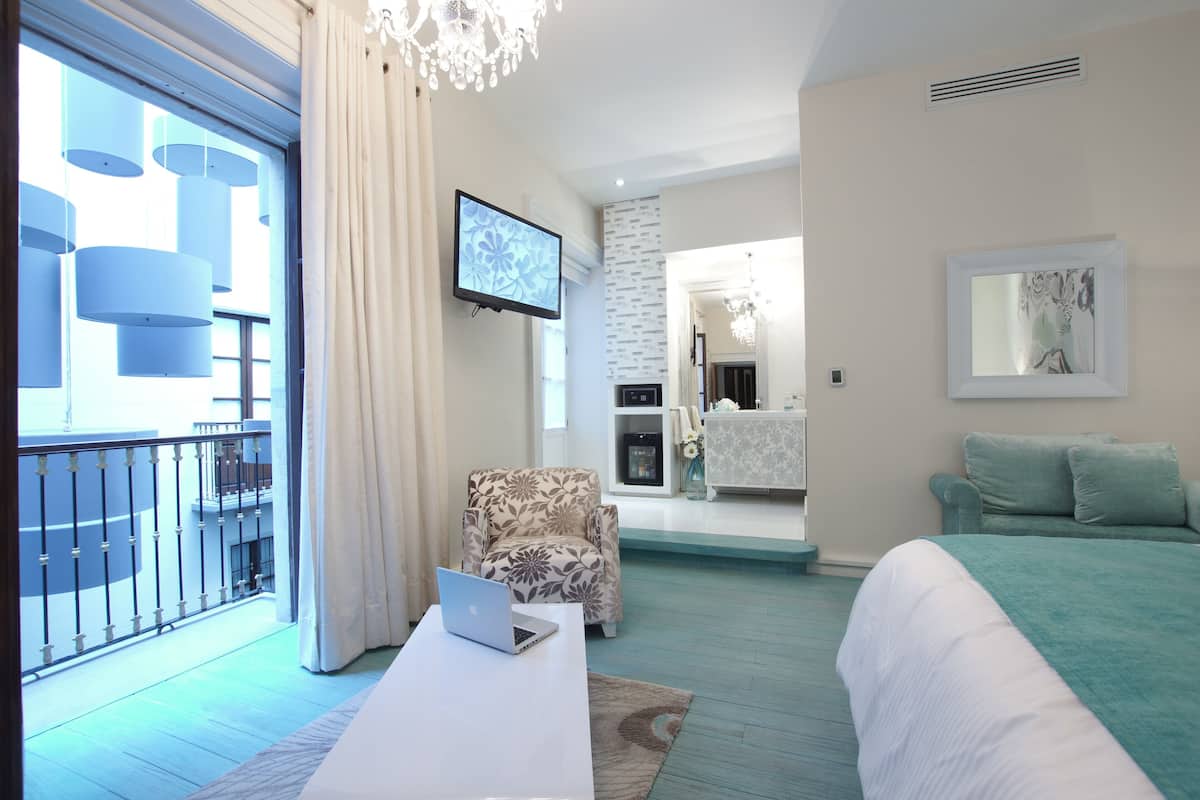 Una habitación de hotel moderna con balcón, cama, escritorio con computadora portátil, televisor y baño con espejo y lavamanos.