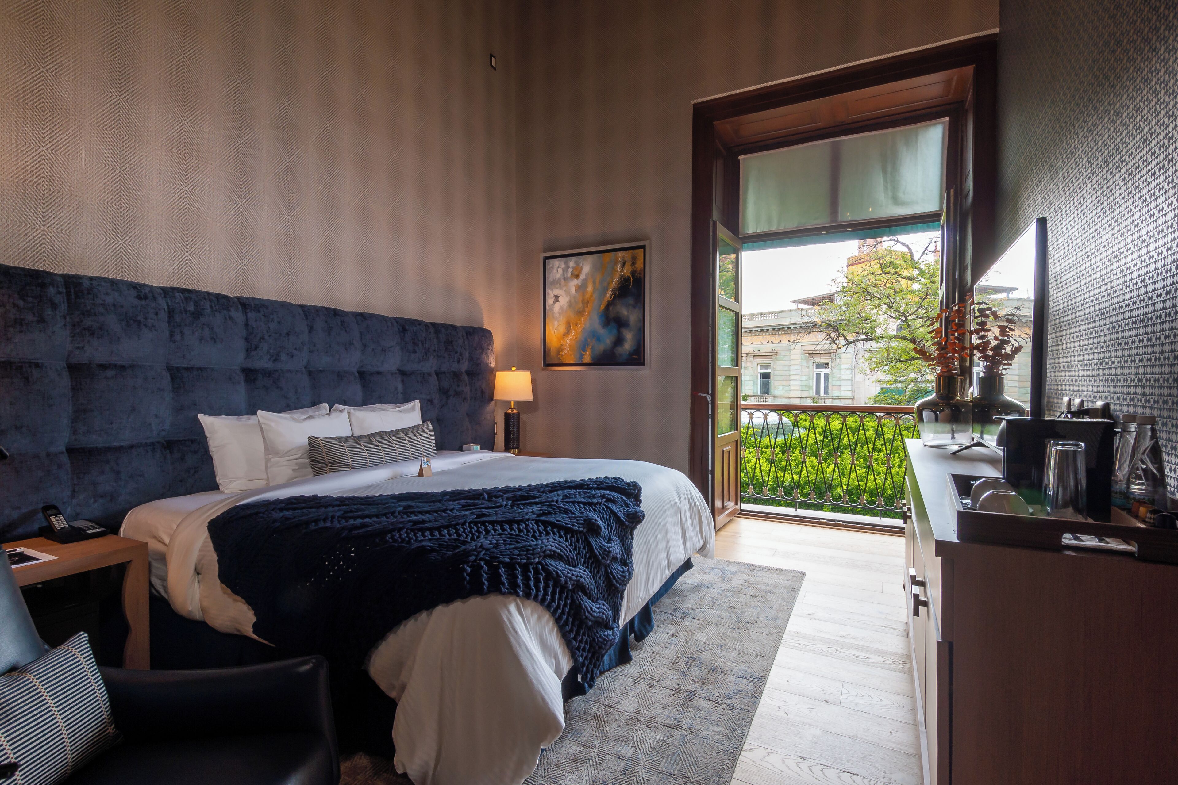 Una habitación de hotel moderna con una cama grande, un escritorio, una silla, una mesita de noche, una lámpara, un cuadro y un balcón con vista a la ciudad.