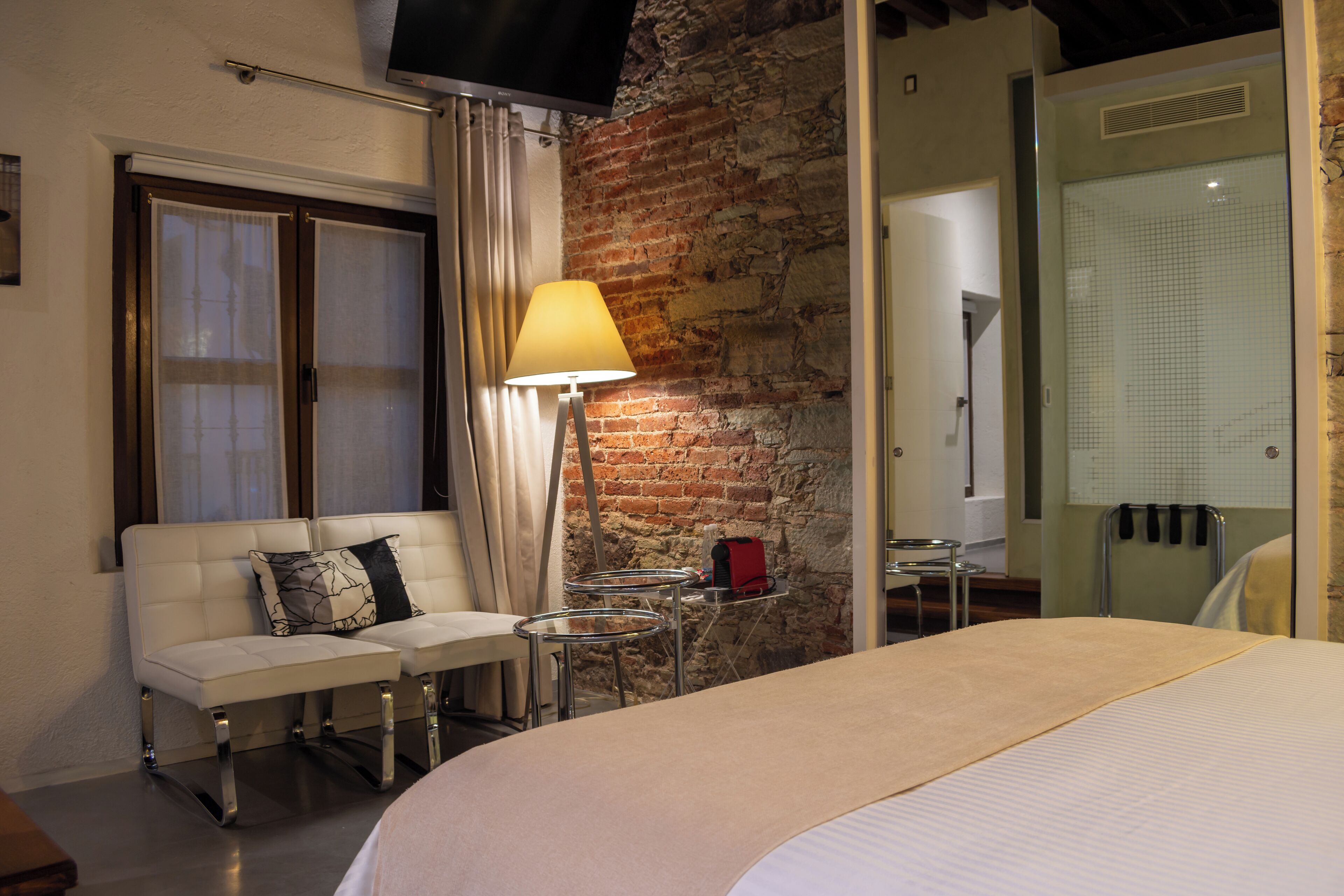 A hotel room with a brick wall, a sofa with cushions, a bedside table with a lamp, and a bed with a white comforter.