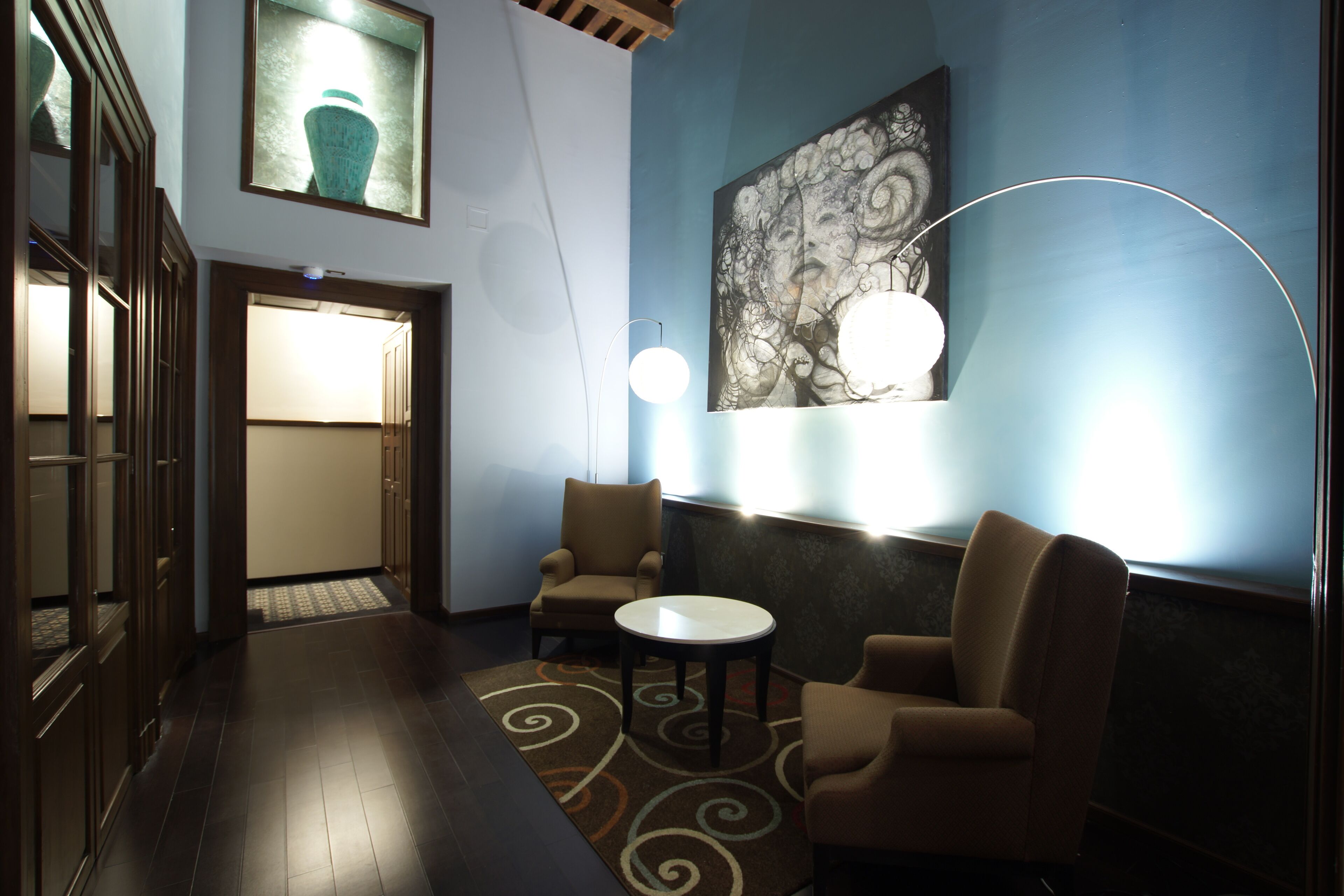 Un vestíbulo con suelo de madera, dos sillones, una mesita redonda, una lámpara de pared y un cuadro abstracto de gran tamaño.
