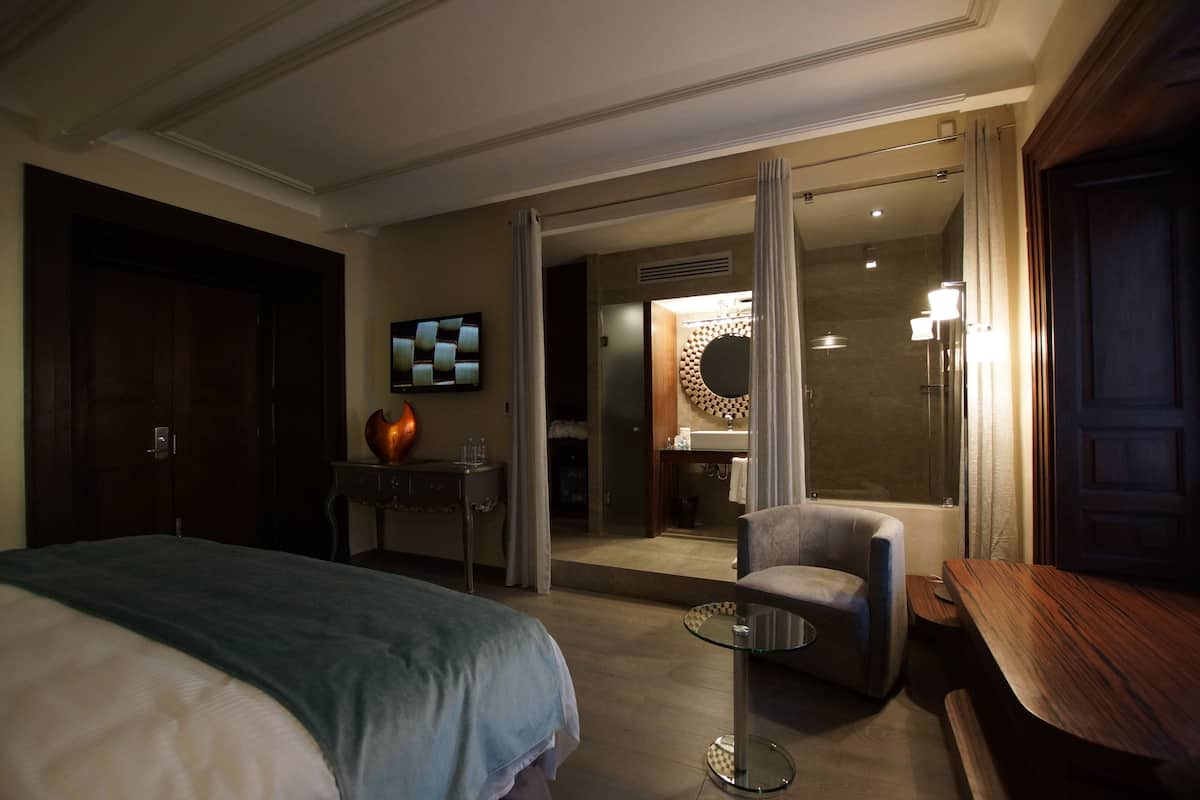 Habitación de hotel con cama, una silla, una mesita, televisor y baño visible a través de una puerta abierta.