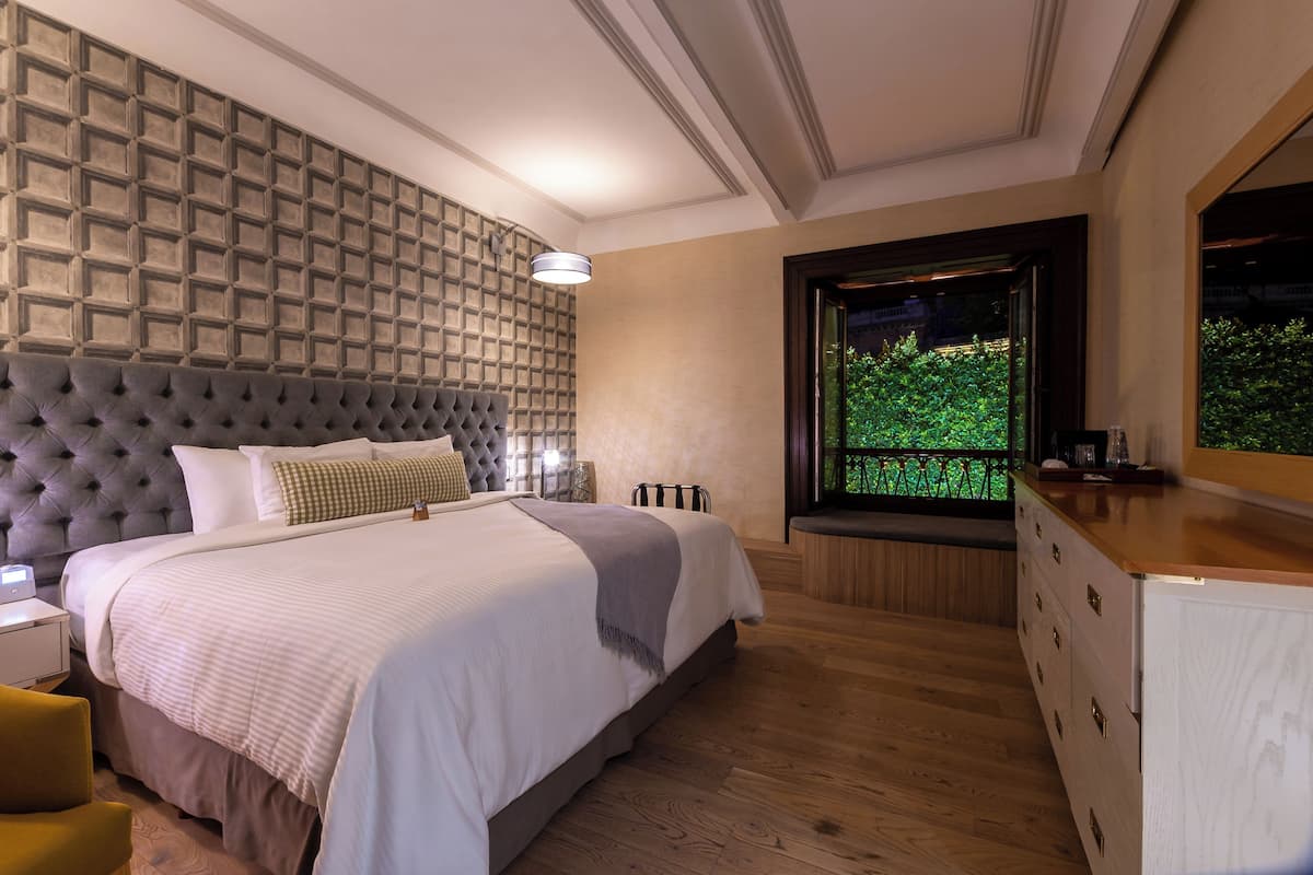 Un dormitorio con cabecera capitonada, una cama grande con ropa de cama blanca, un aparador de madera y una ventana con vistas a zonas verdes.