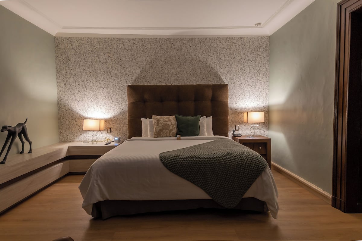 Una cama bien hecha con cabecera marrón, dos lámparas de noche y una pared decorativa con textura.