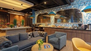 A modern hotel lobby with a sofa, armchairs, a coffee station, and a decorative wall.