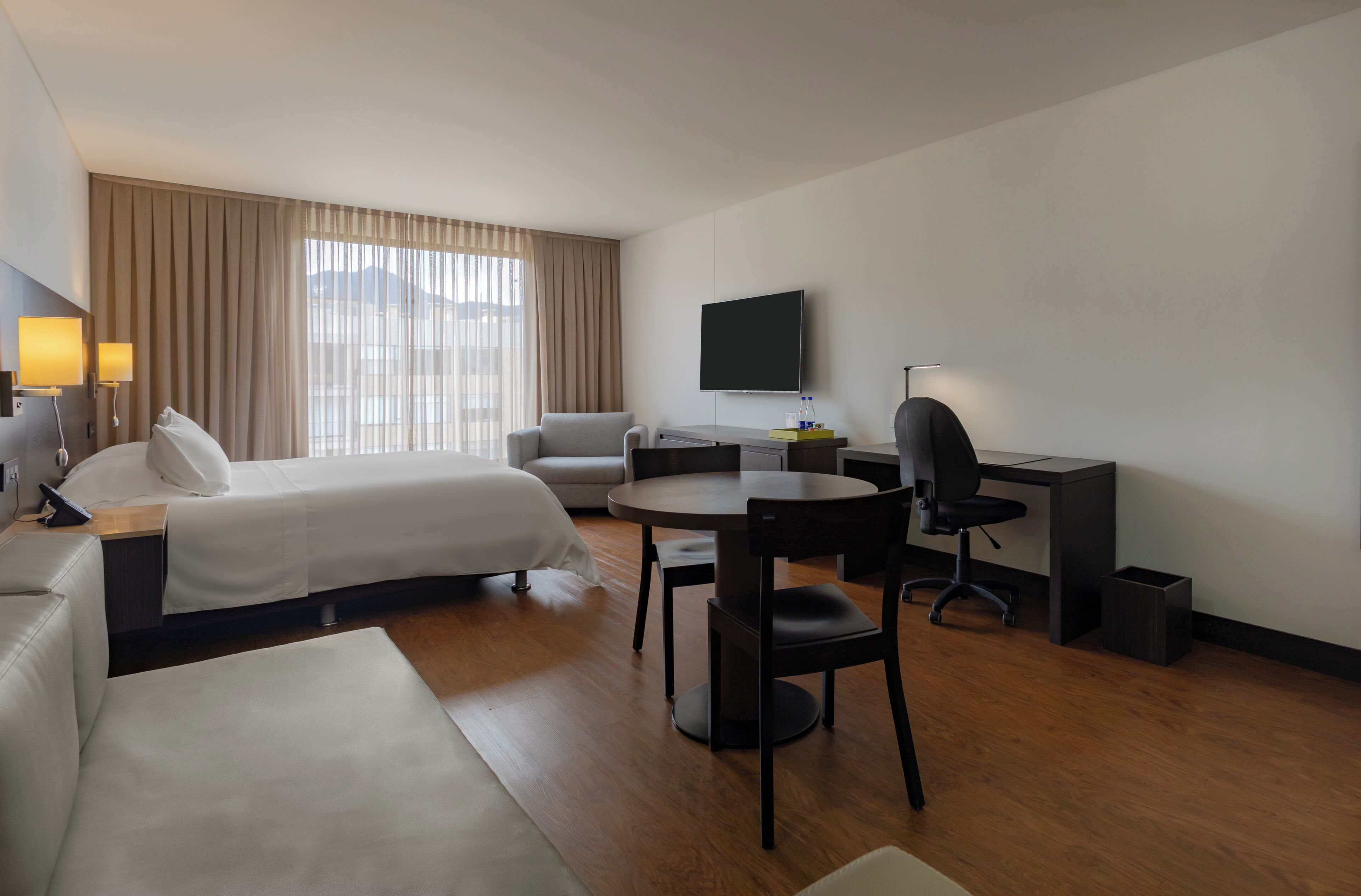 Una habitación de hotel con cama, escritorio, silla, televisor y ventana con cortinas.