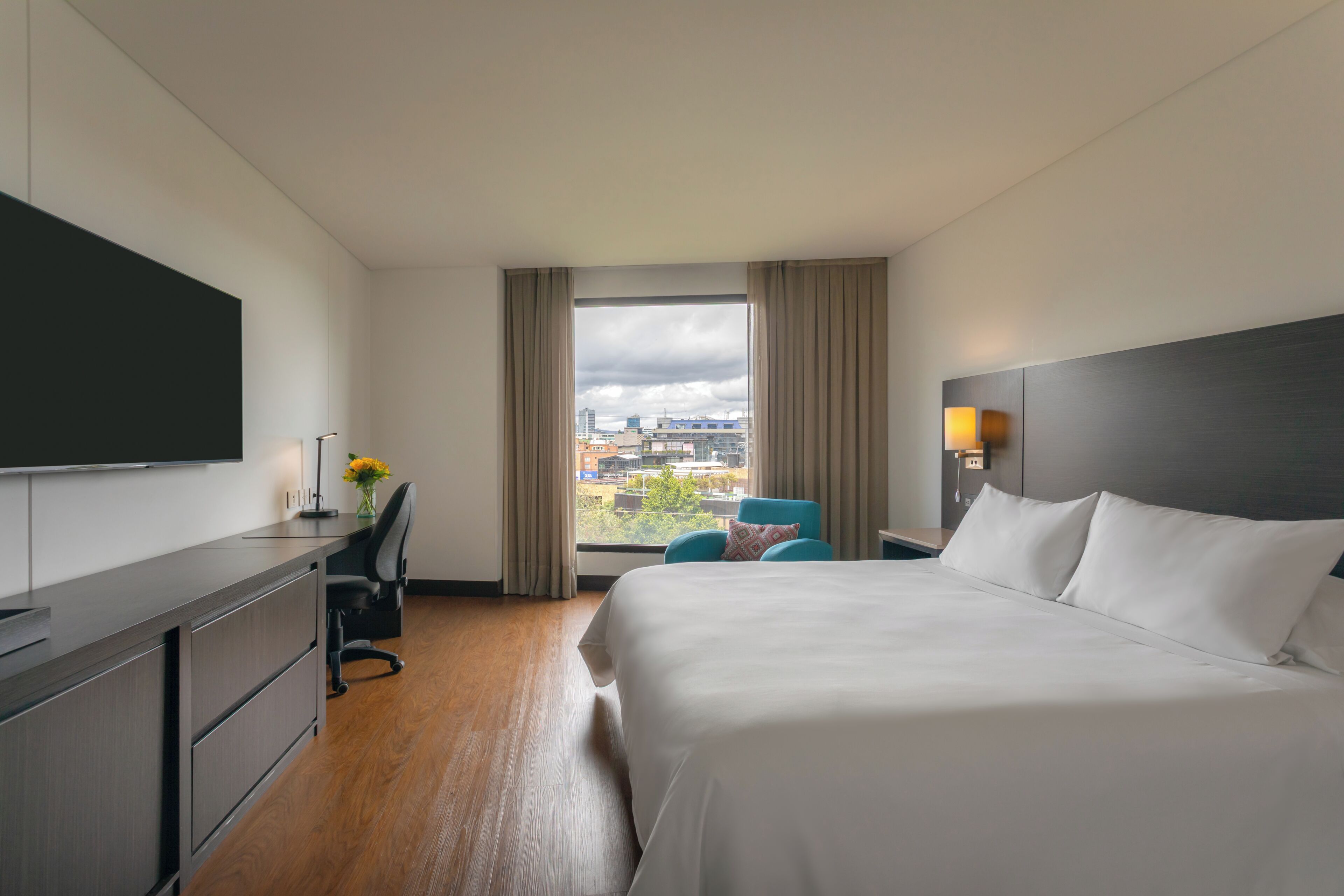 A hotel room with a bed, desk, chair, TV, and a window with a city view.