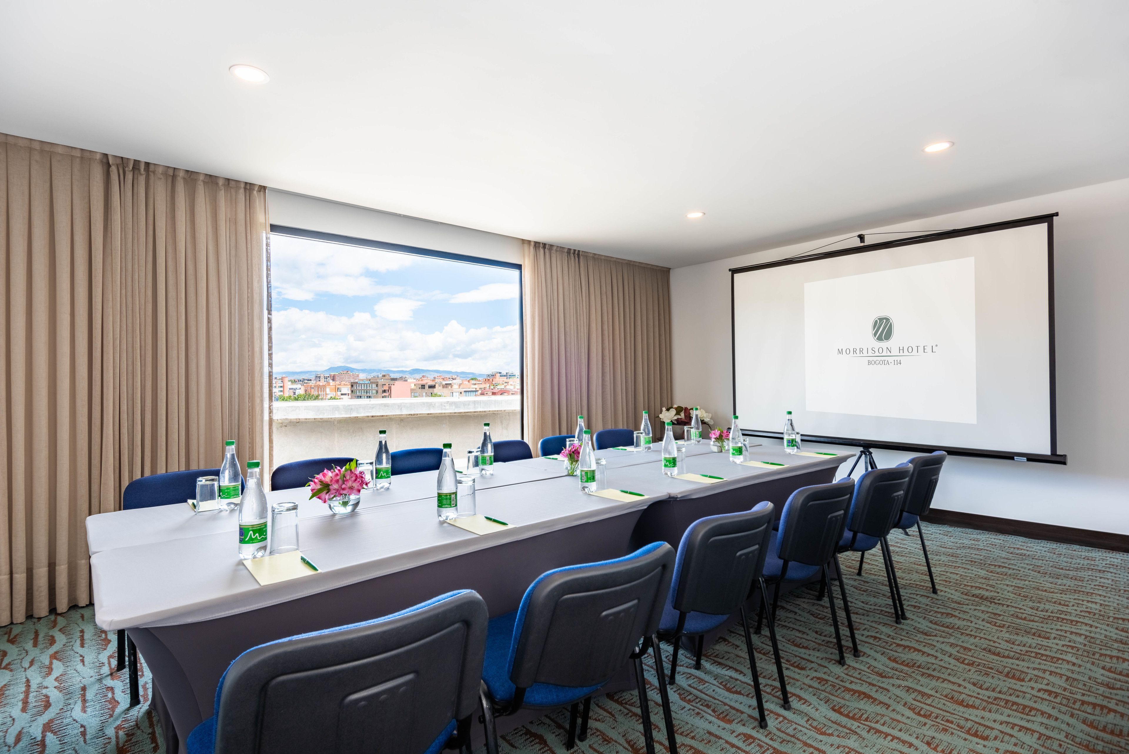 A conference room with a long table, chairs, a projector screen, and a view of the city through large windows.