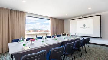 A conference room with a long table, chairs, a projector screen, and a view of the city through large windows.