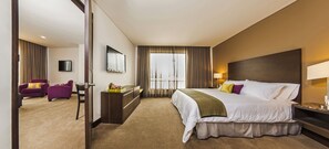Una habitación de hotel con una cama grande, un escritorio, dos sillones morados, un televisor y una ventana con cortinas.