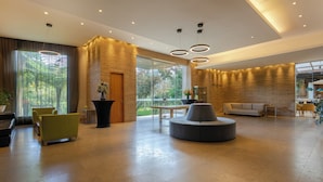 A spacious lobby with a modern design, featuring a round seating area, a large potted plant, and a view through a glass door.