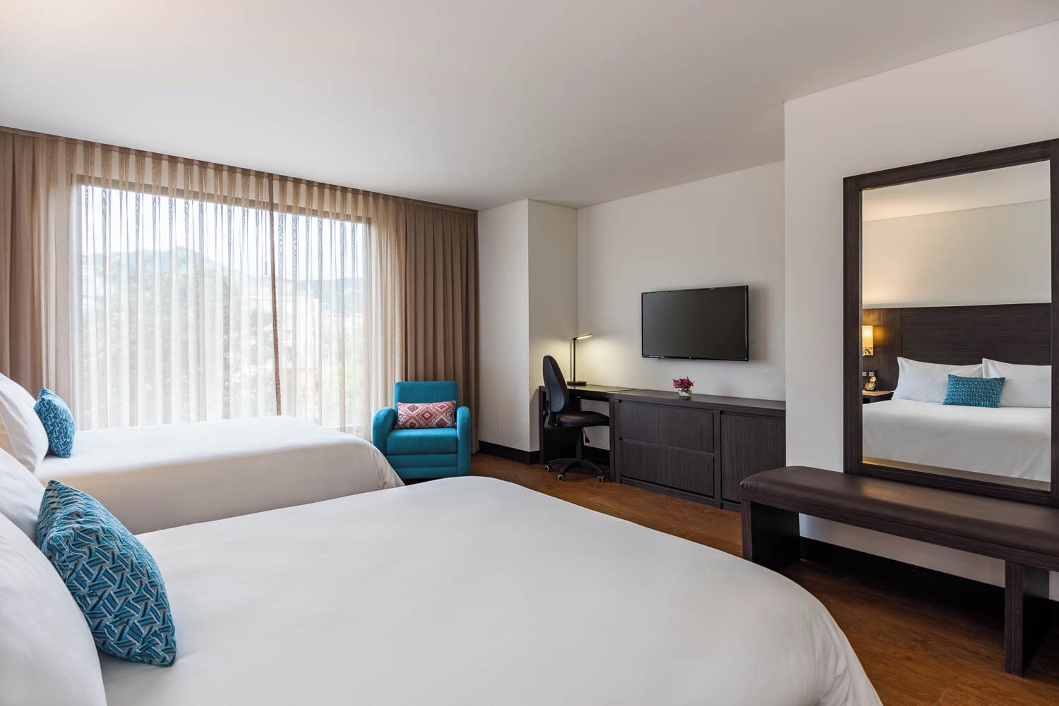 Habitación de hotel con dos camas, un escritorio, una silla, un televisor y un espejo grande.