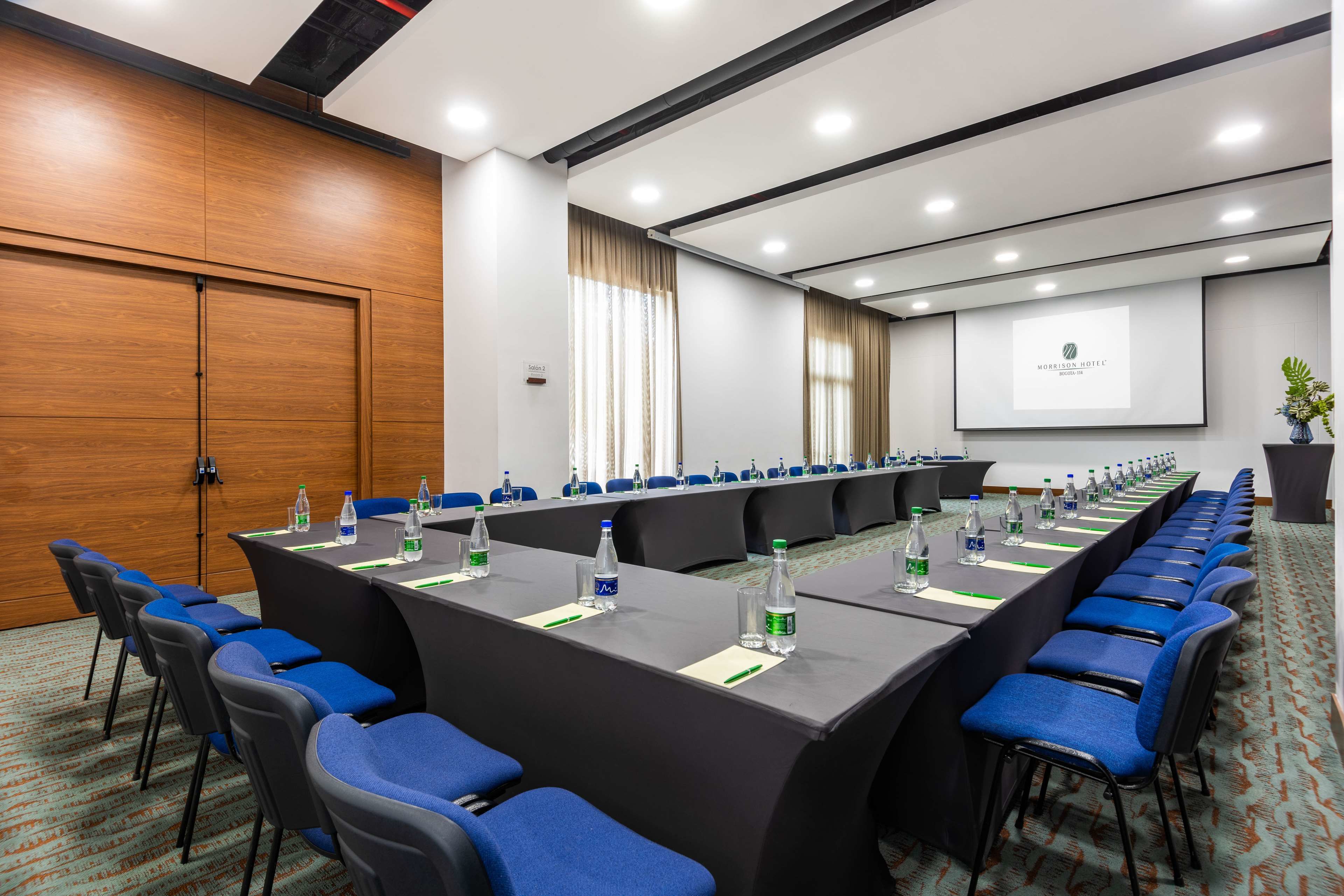 A conference room with a long table, blue chairs, water bottles, and a projector screen displaying the hotel name.