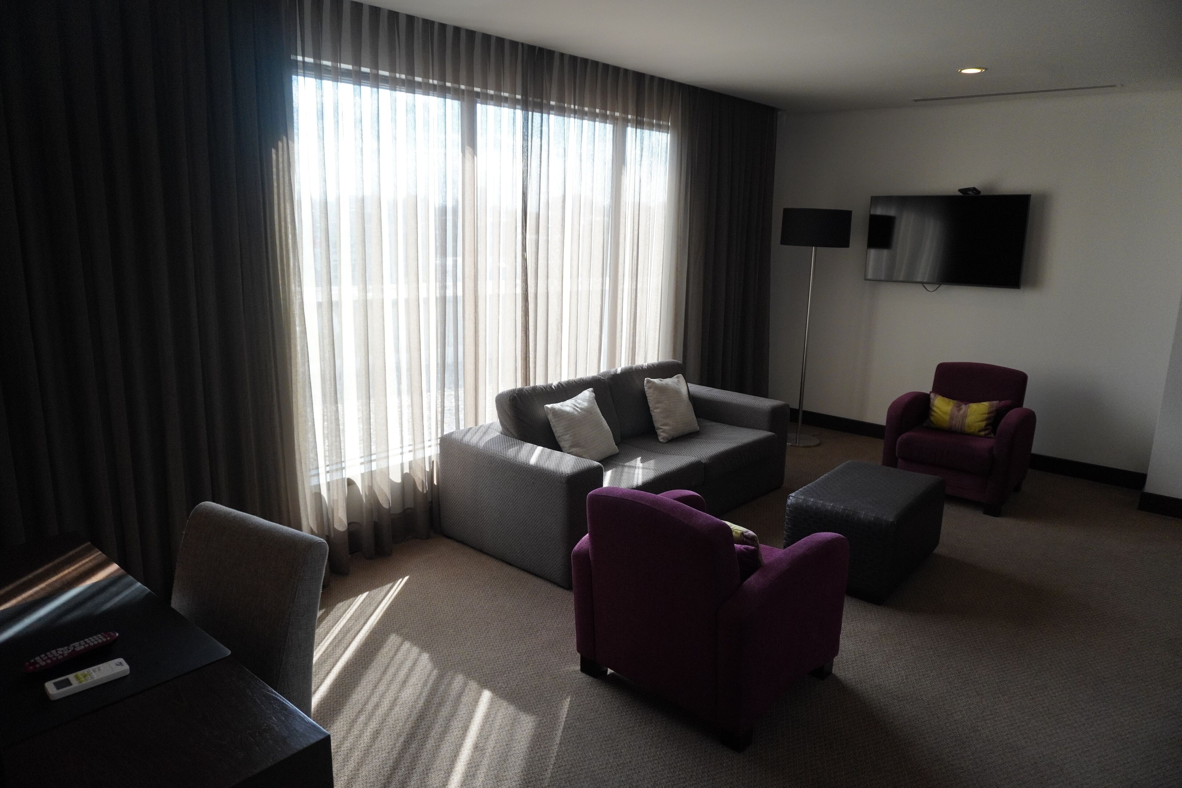 Moderni hotellihuone, jossa on harmaa sohva, kaksi violettia nojatuolia, taulutelevisio ja suuri ikkuna, jossa on läpikuultavat verhot.
