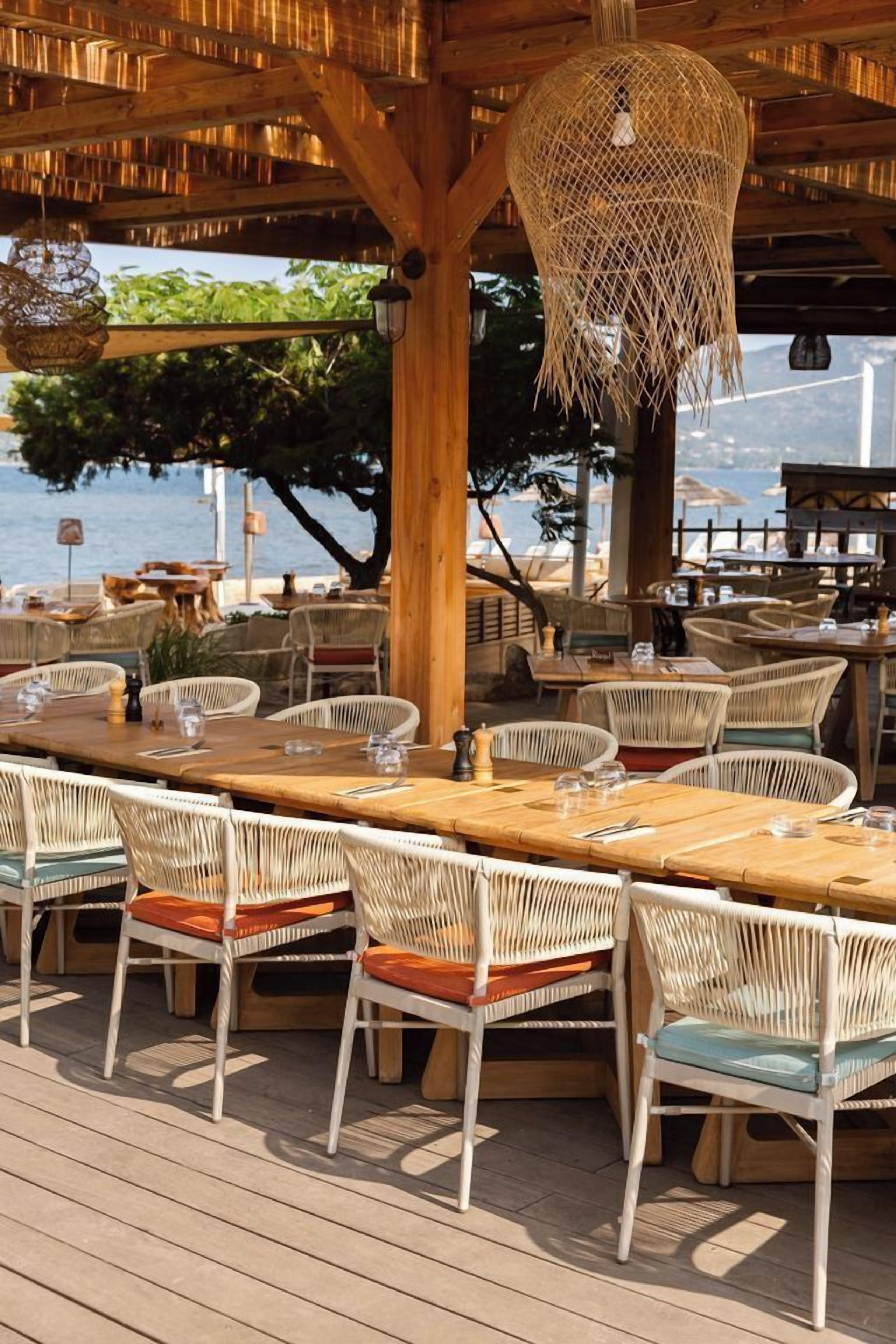 An outdoor dining area with wooden tables and wicker chairs, overlooking a body of water.