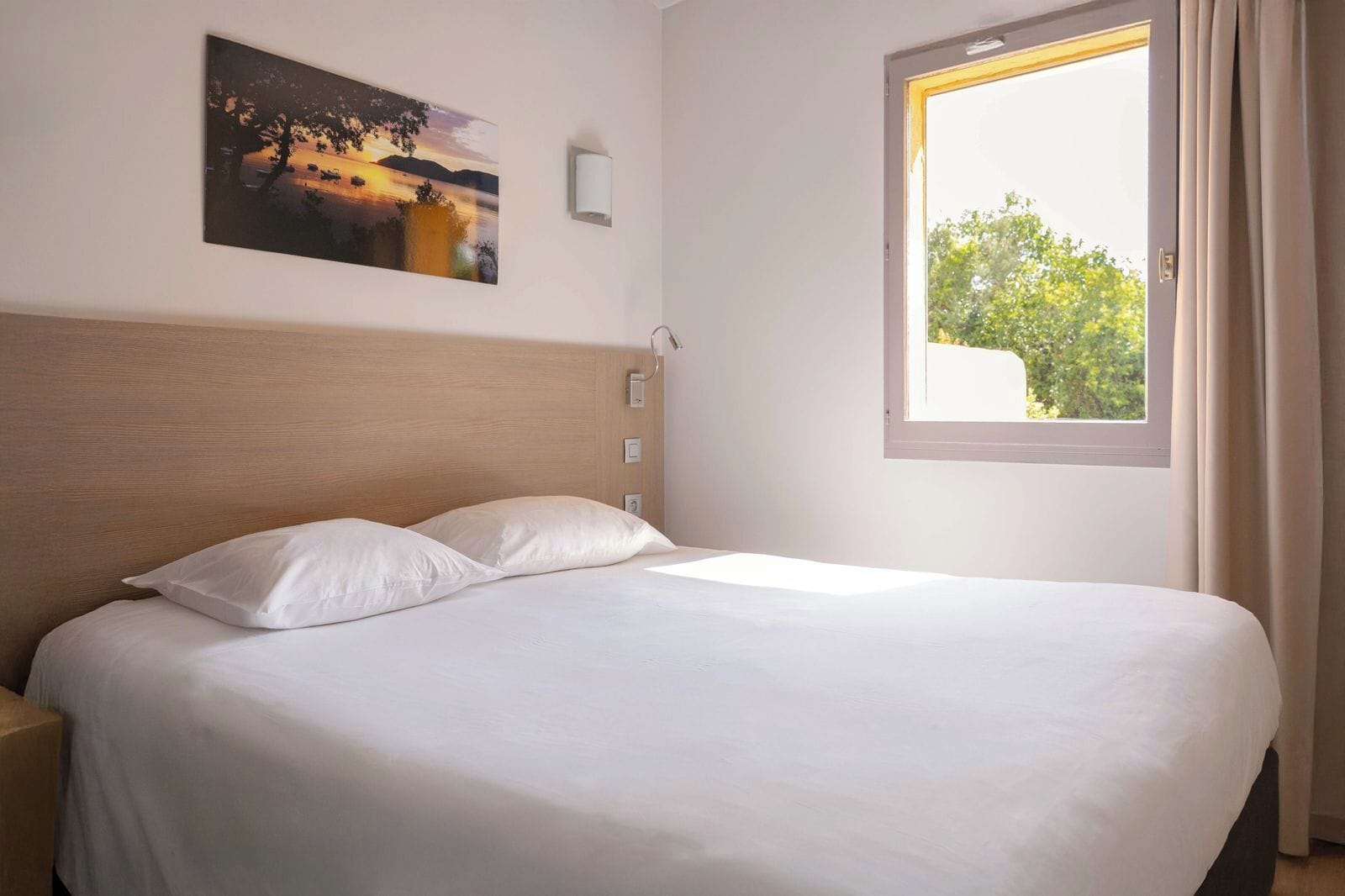 A neatly made bed with white linens in a room with a wooden headboard, a painting on the wall, and a window with curtains.
