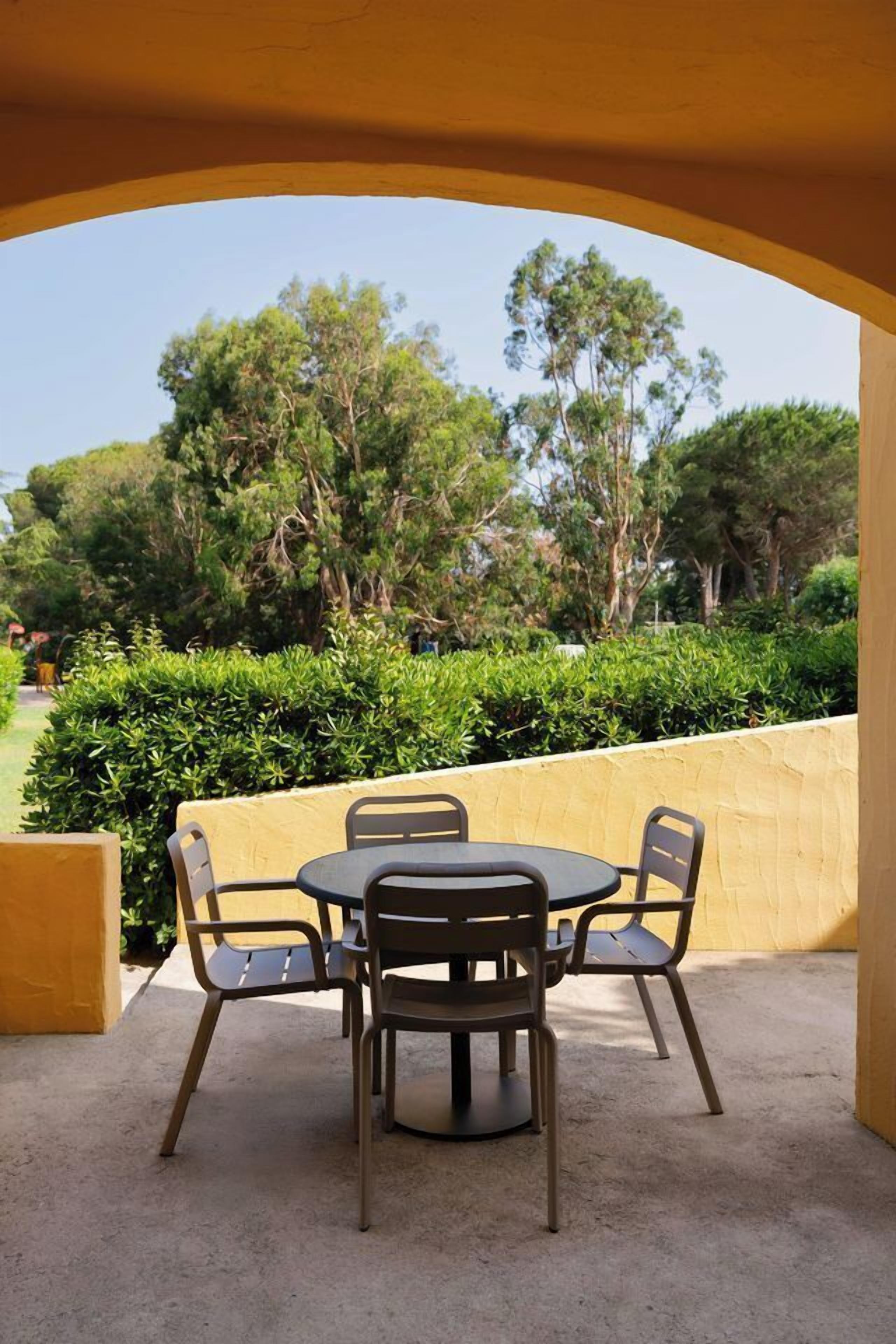 Een overdekt terras met een ronde tafel en vier stoelen, omringd door een gele muur en uitzicht op bomen en groen.