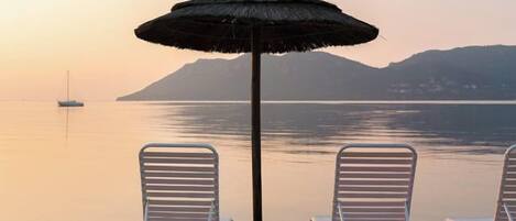 A beach scene with two white lounge chairs and a thatched umbrella.