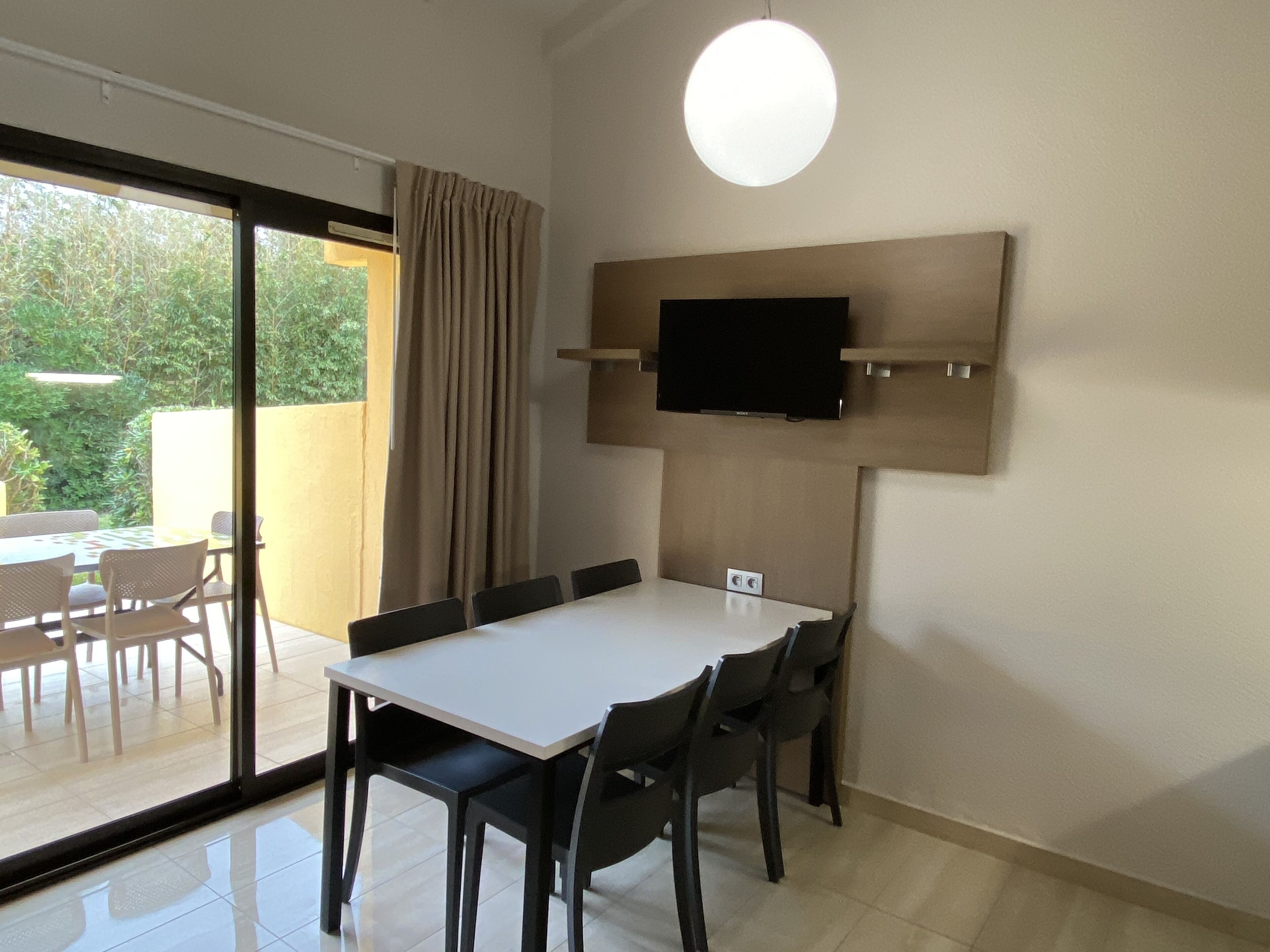 A modern room with a large window, a dining table with chairs, a flat-screen TV mounted on the wall, and a sliding glass door leading to an outdoor area with a table and chairs.