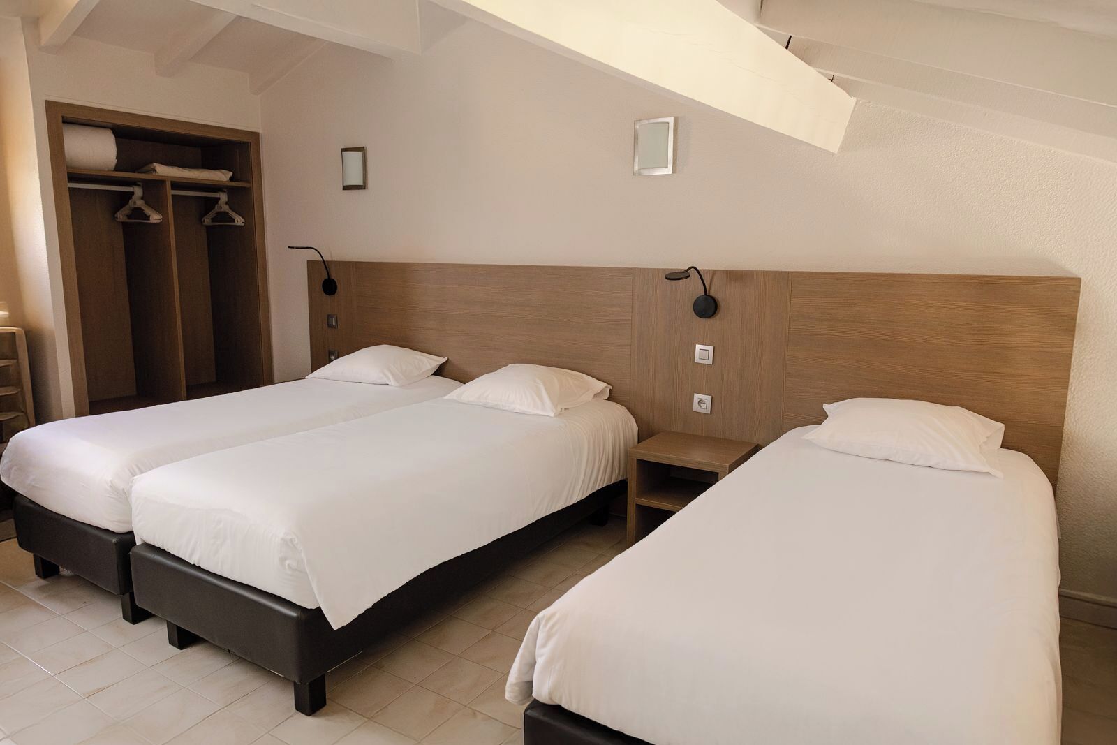 A hotel room with two single beds, a wooden headboard, a small bedside table, and a wardrobe with hanging clothes.