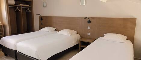 A hotel room with two single beds, a wooden headboard, a small bedside table, and a wardrobe with hanging clothes.