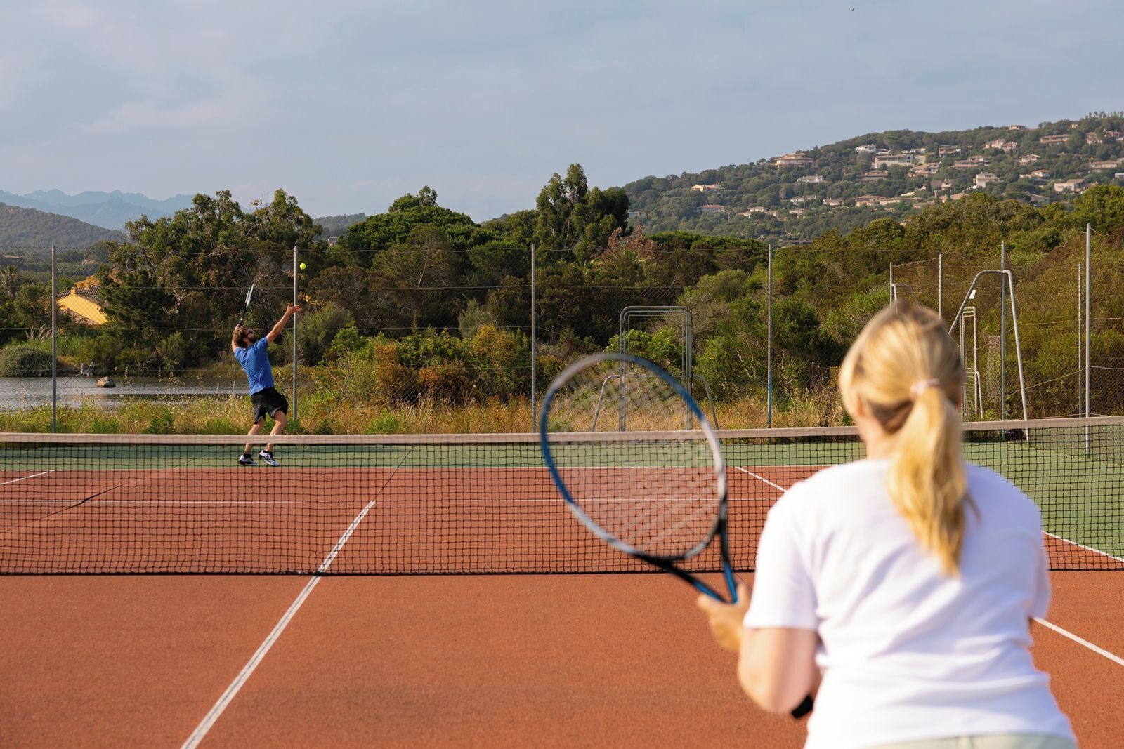 Una cancha de tenis con red, rodeada de vegetación y un paisaje montañoso al fondo.