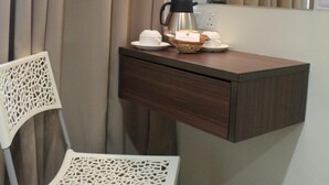 Un pequeño escritorio con una cafetera, una cesta y un cubo de basura en una habitación con una silla y cortinas.