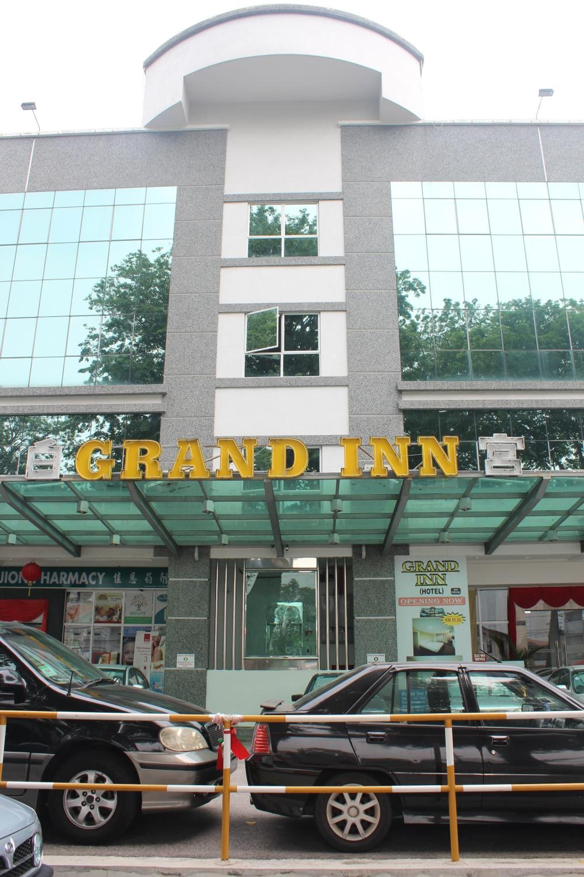 La façade de l’hôtel Grand Inn, avec son enseigne bien visible, son entrée en verre et, au fond, une pharmacie dont l’enseigne est également visible.
