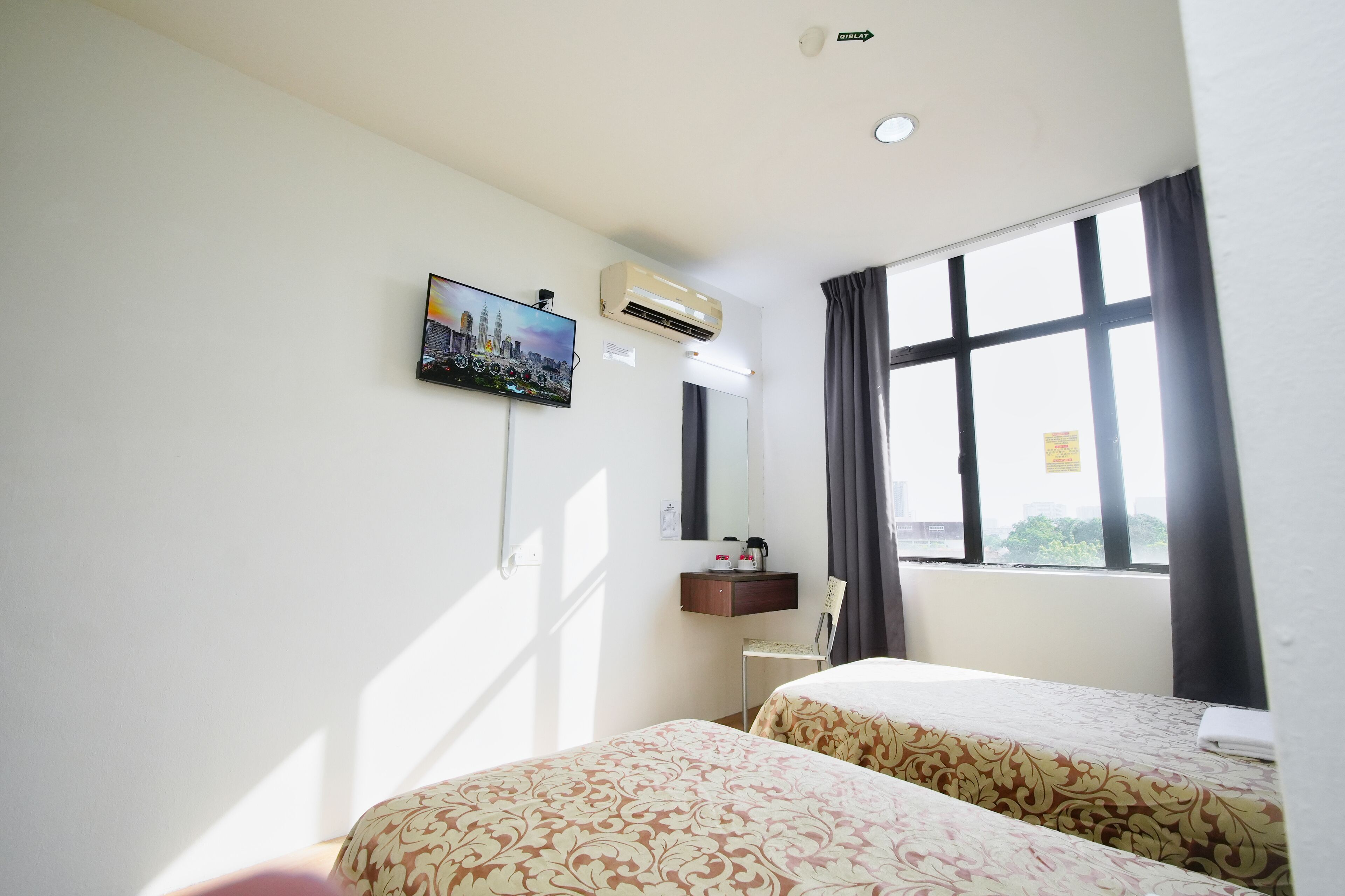 A hotel room with two beds, a TV mounted on the wall, an air conditioning unit, a small desk with a chair, and a window with curtains.