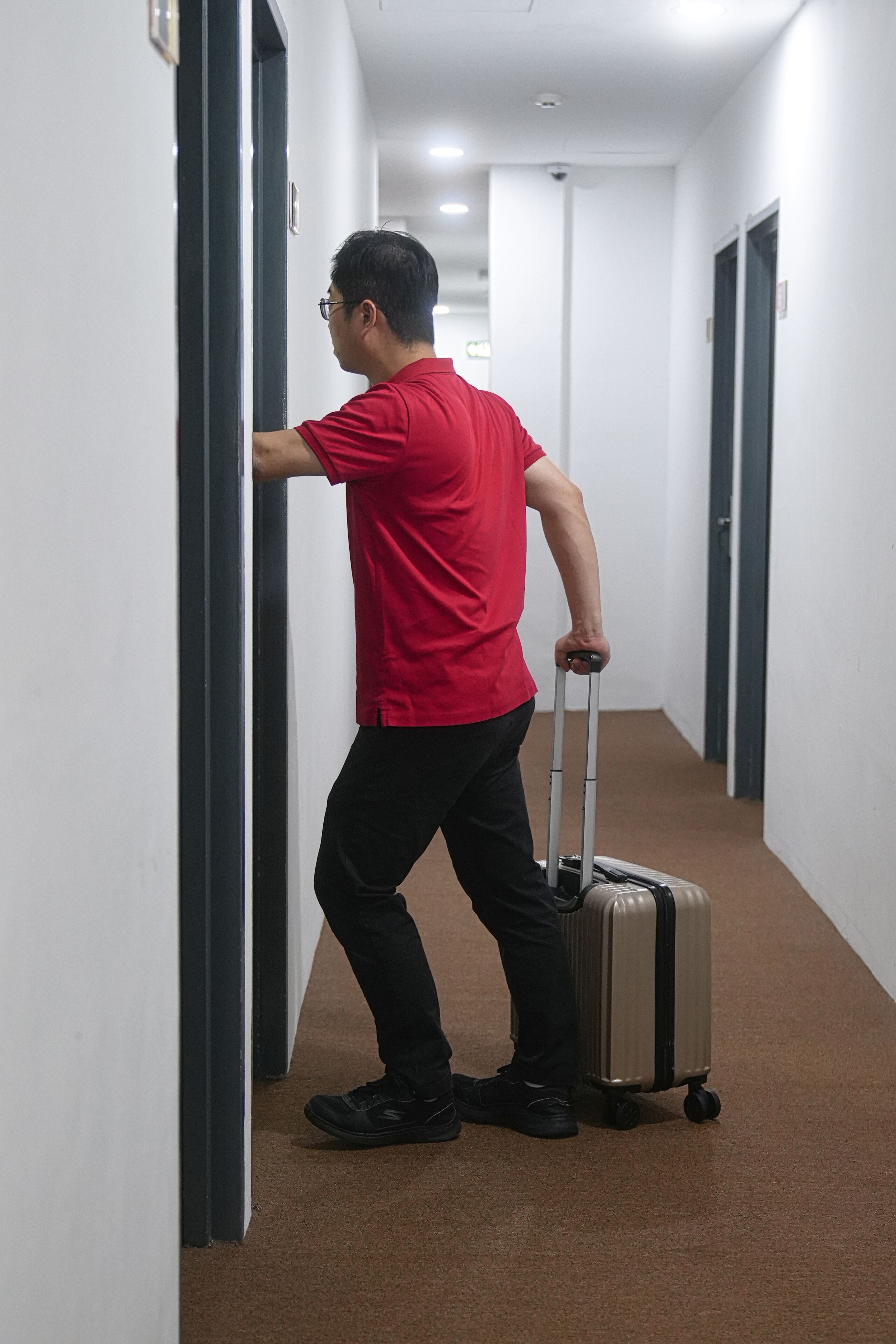 A man in a red shirt pushing a suitcase down a hallway with white walls and a brown carpet.