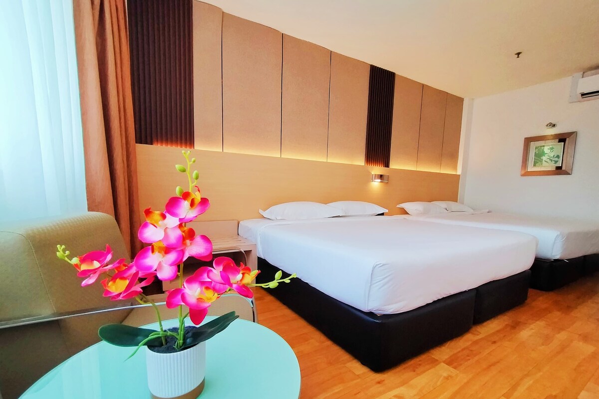 Una habitación de hotel con cama, mesitas de noche, una orquídea en maceta, una silla y una pequeña mesa.