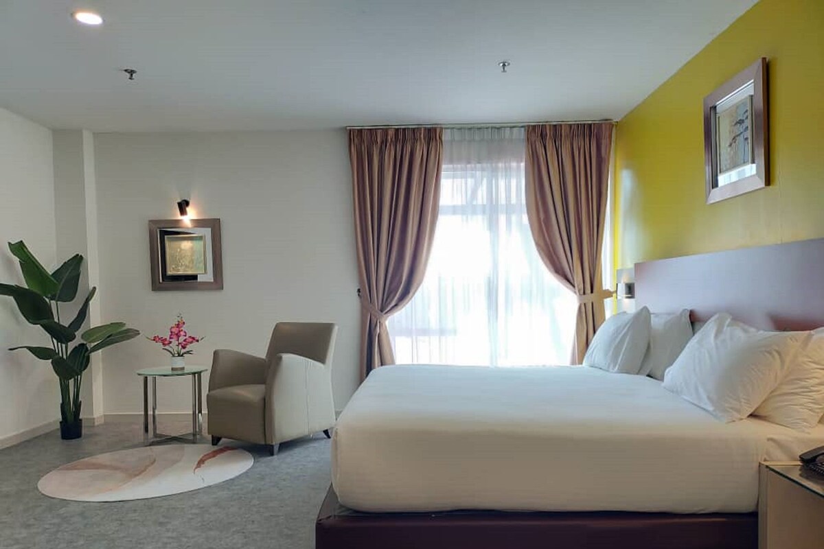 Habitación de hotel con cama, una silla, una mesita con una planta y un cuadro en la pared.