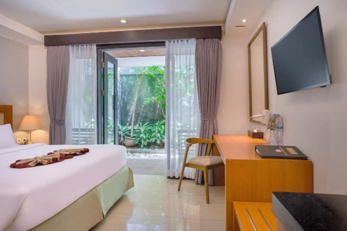 A hotel room with a bed, a desk, a chair, a TV, and a view of greenery outside.