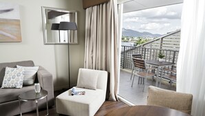 A modern living room with a sofa, armchair, and a dining table with a bowl of apples. A balcony with a view of the city and mountains is visible through large windows. - Grand Hotel and Apartments Townsville (South Townsville)